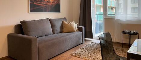 Een moderne woonkamer met een grijze bank, een stadsgezicht schilderij, een glazen tafel, en uitzicht op een balkon met kleurrijke gebouwen.