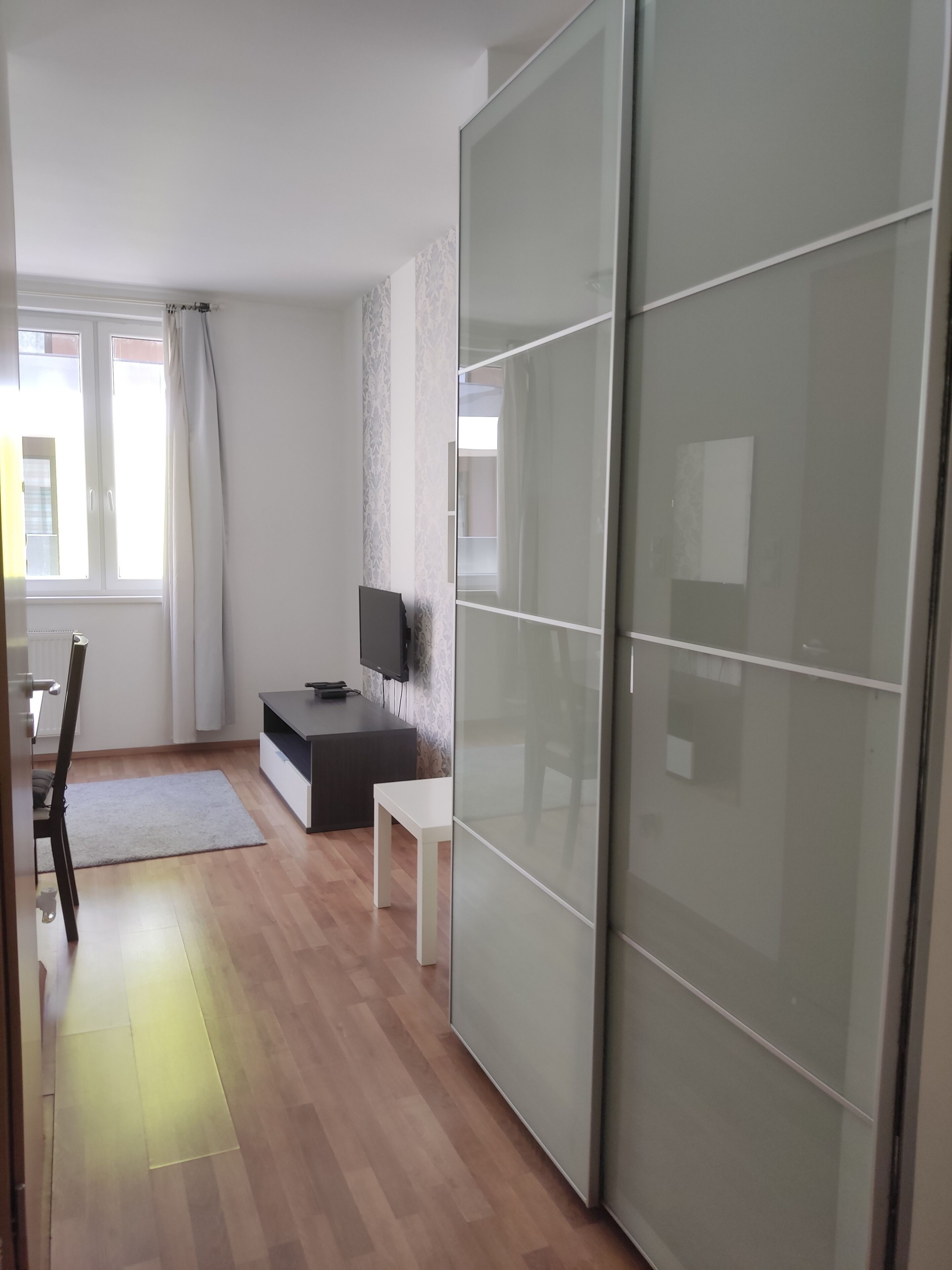 A modern room with a sliding glass door closet, a flat-screen TV on a stand, a small white table, and a wooden floor.