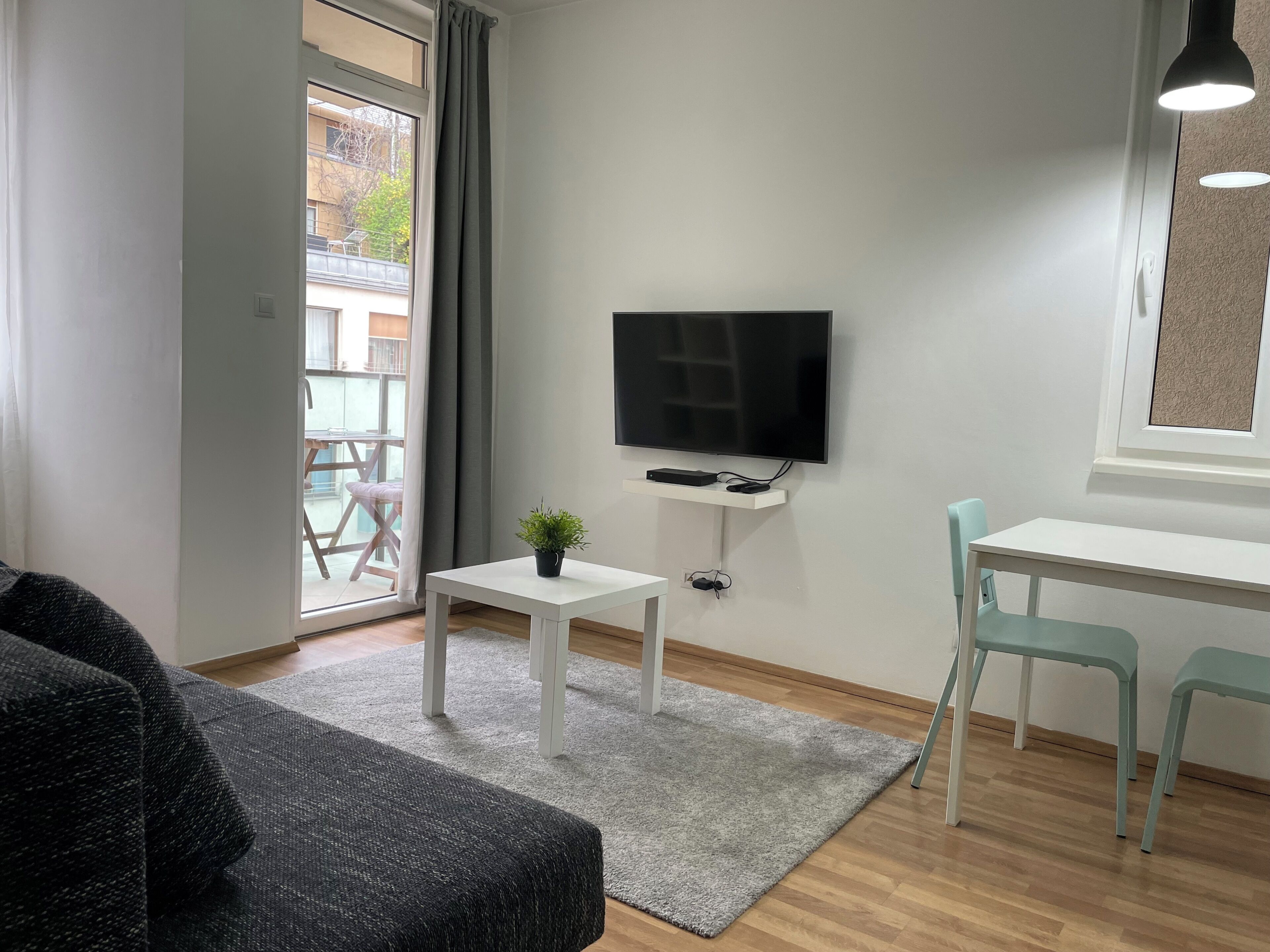 A modern living room with a flat-screen TV, a small table with a plant, a sofa, and a desk with two chairs.