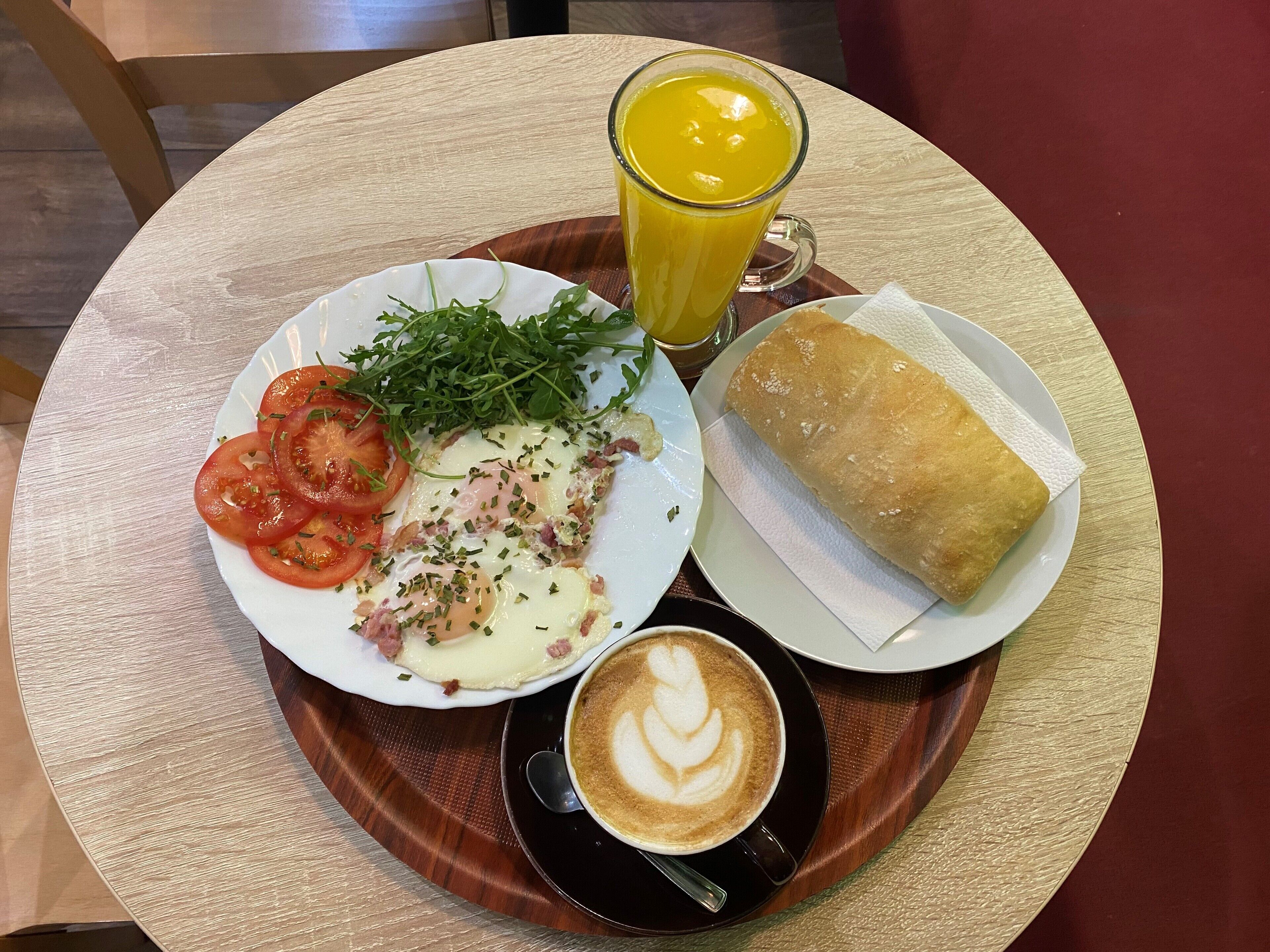 A meal consisting of a plate with tomatoes, greens, and eggs, a glass of orange juice, a piece of bread, and a cup of coffee with latte art.