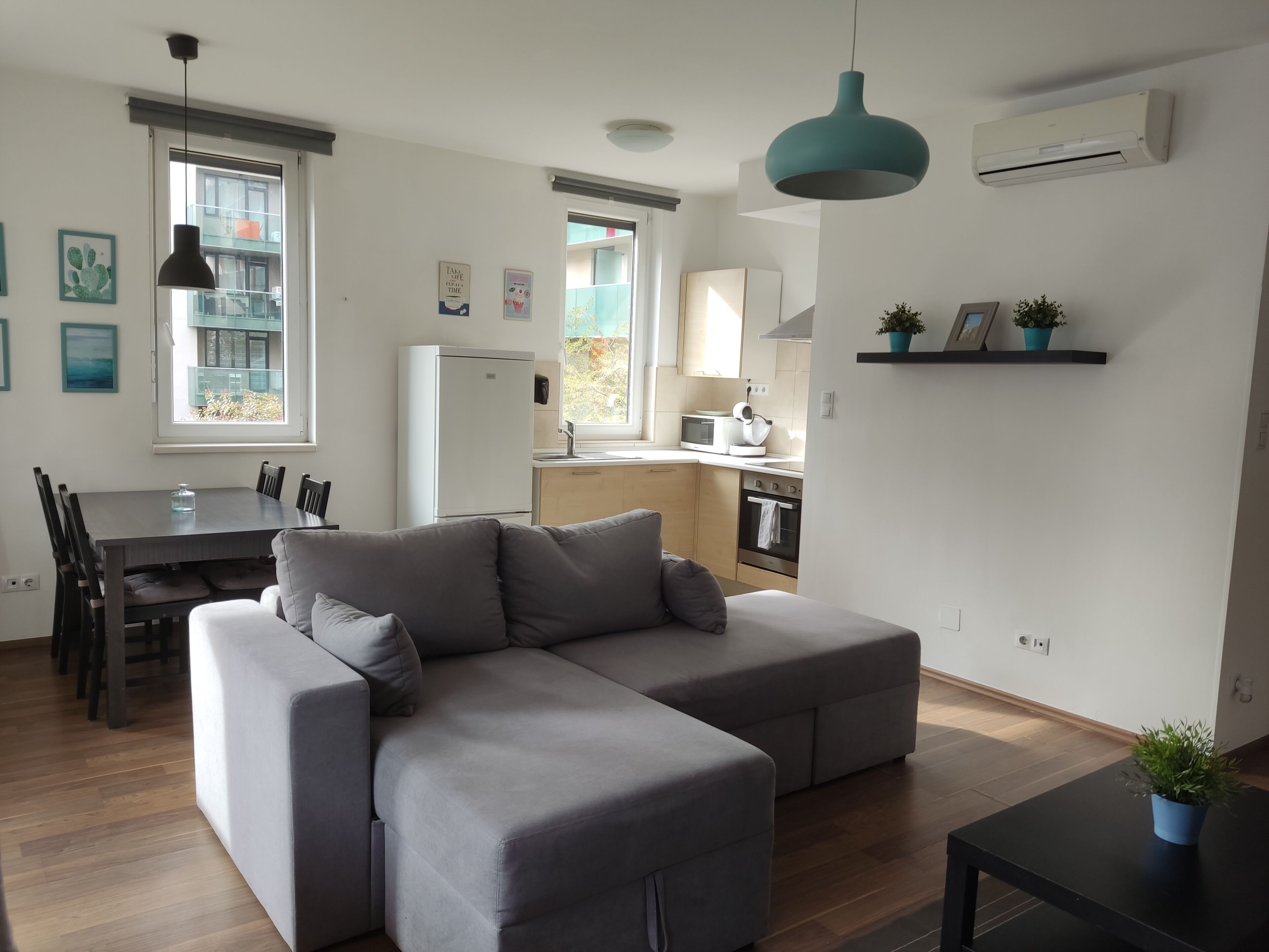 A modern living room with a grey sectional sofa, a dining table with chairs, a kitchen area with a fridge, stove, and microwave, and a window with a view of a balcony.