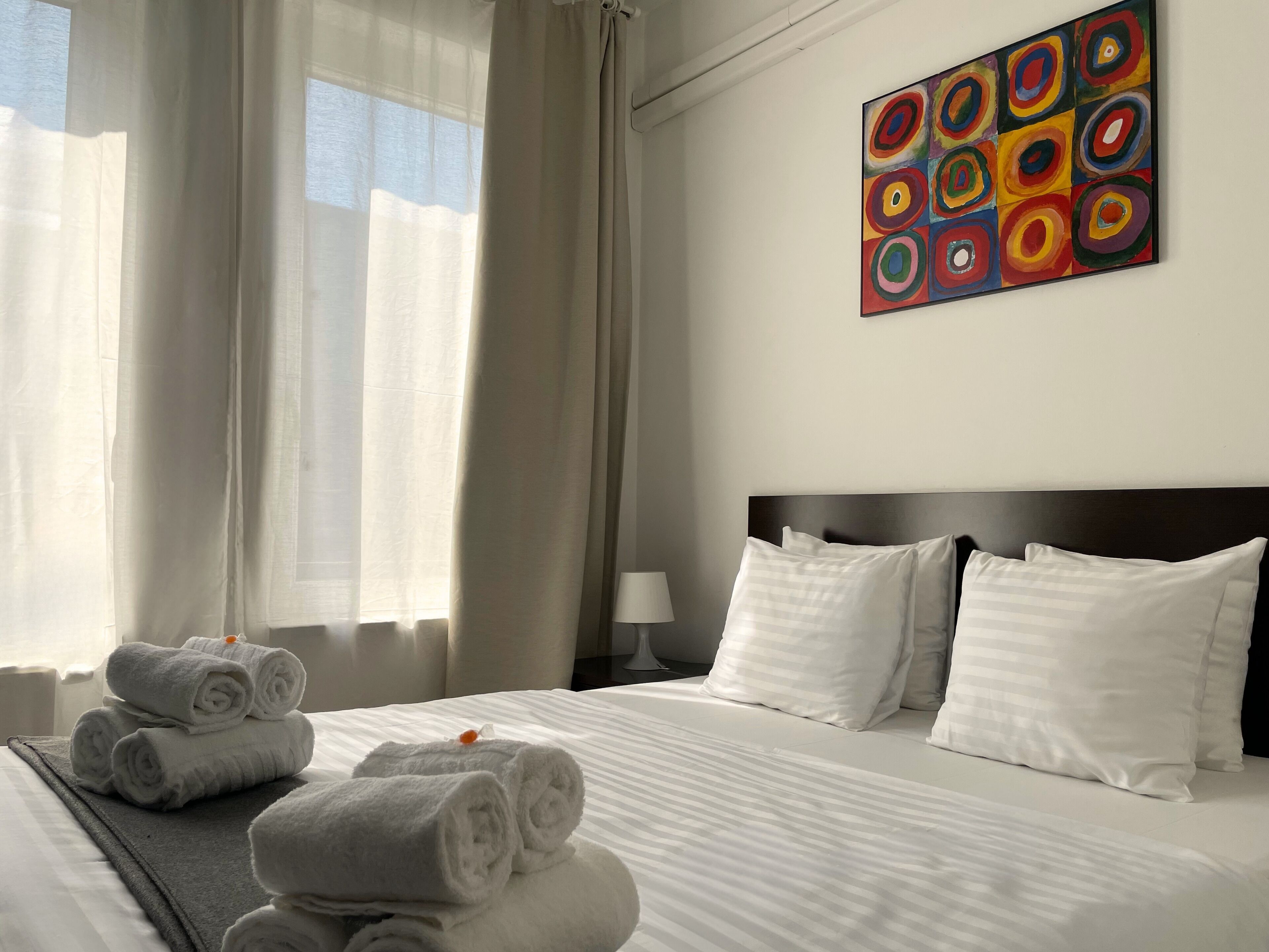 A hotel room with a neatly made bed, a painting on the wall, and a window with curtains.