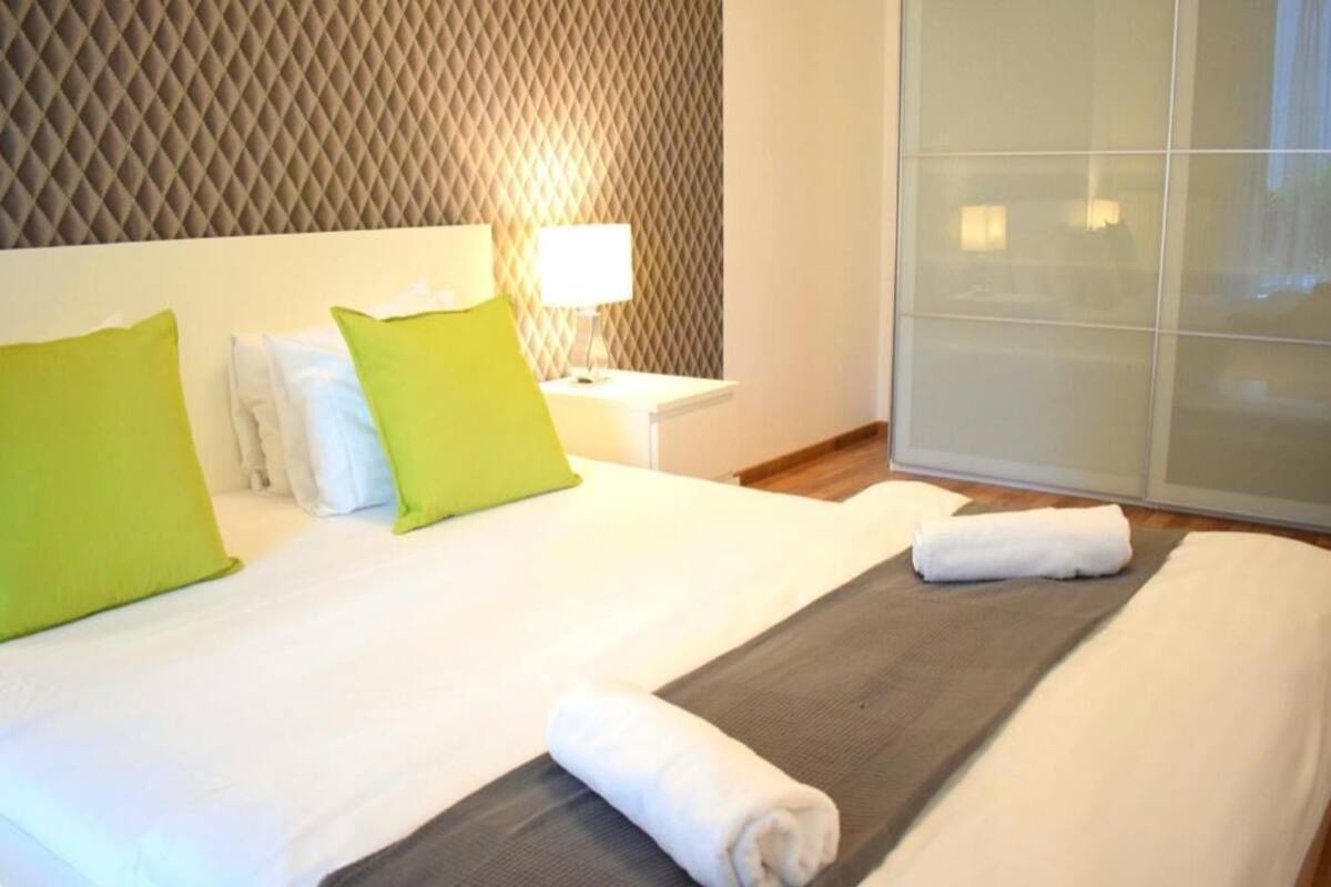 A hotel room with a bed, pillows, a nightstand with a lamp, and a sliding door.