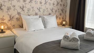 A neatly made bed with white bedding and two pillows, a bedside table with a lamp, and a window with curtains.