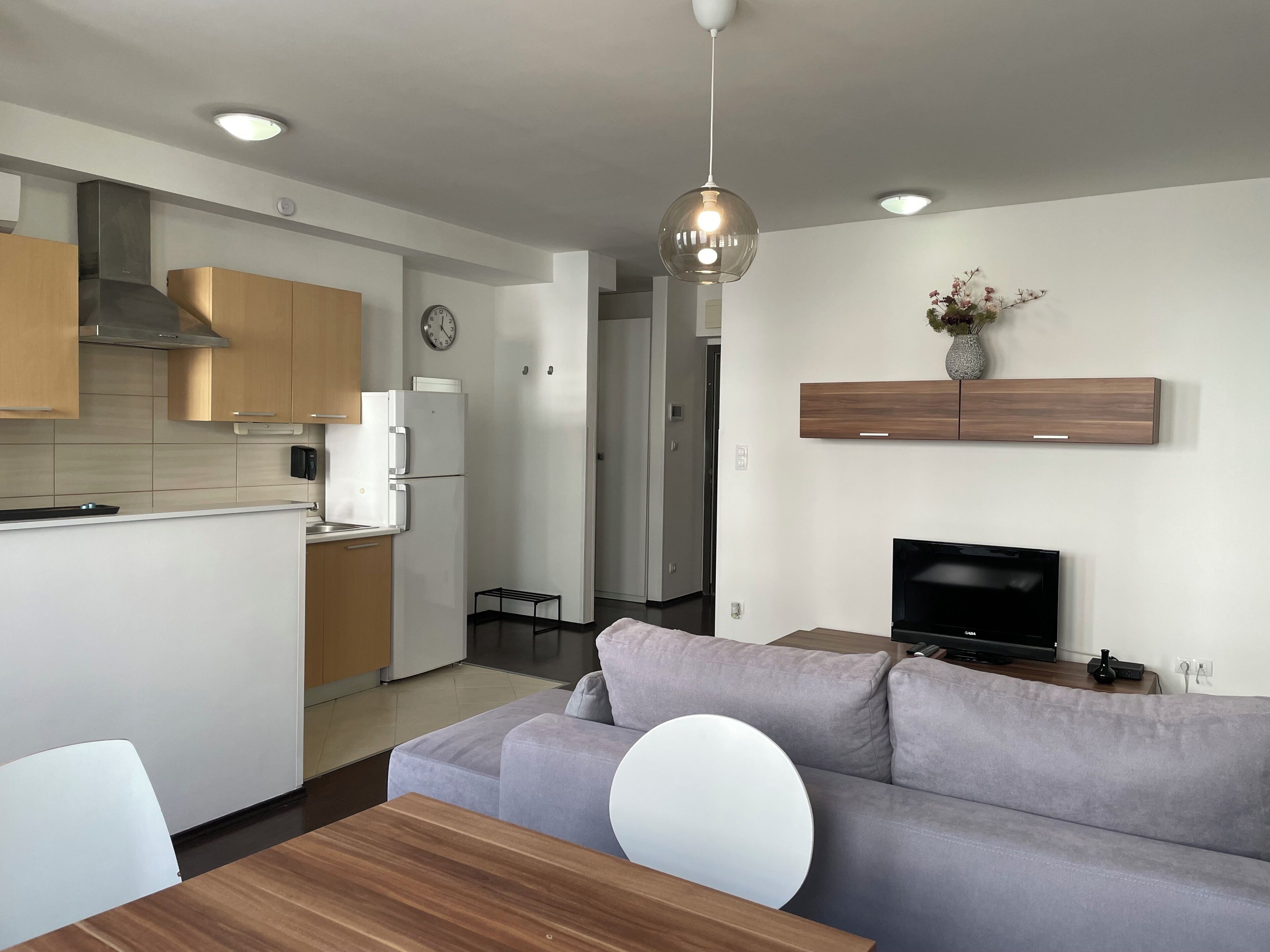 Una cocina moderna con armarios blancos, horno empotrado y microondas. Hay un sofá gris y una mesa de comedor de madera con sillas. Un televisor está montado en la pared sobre el sofá.