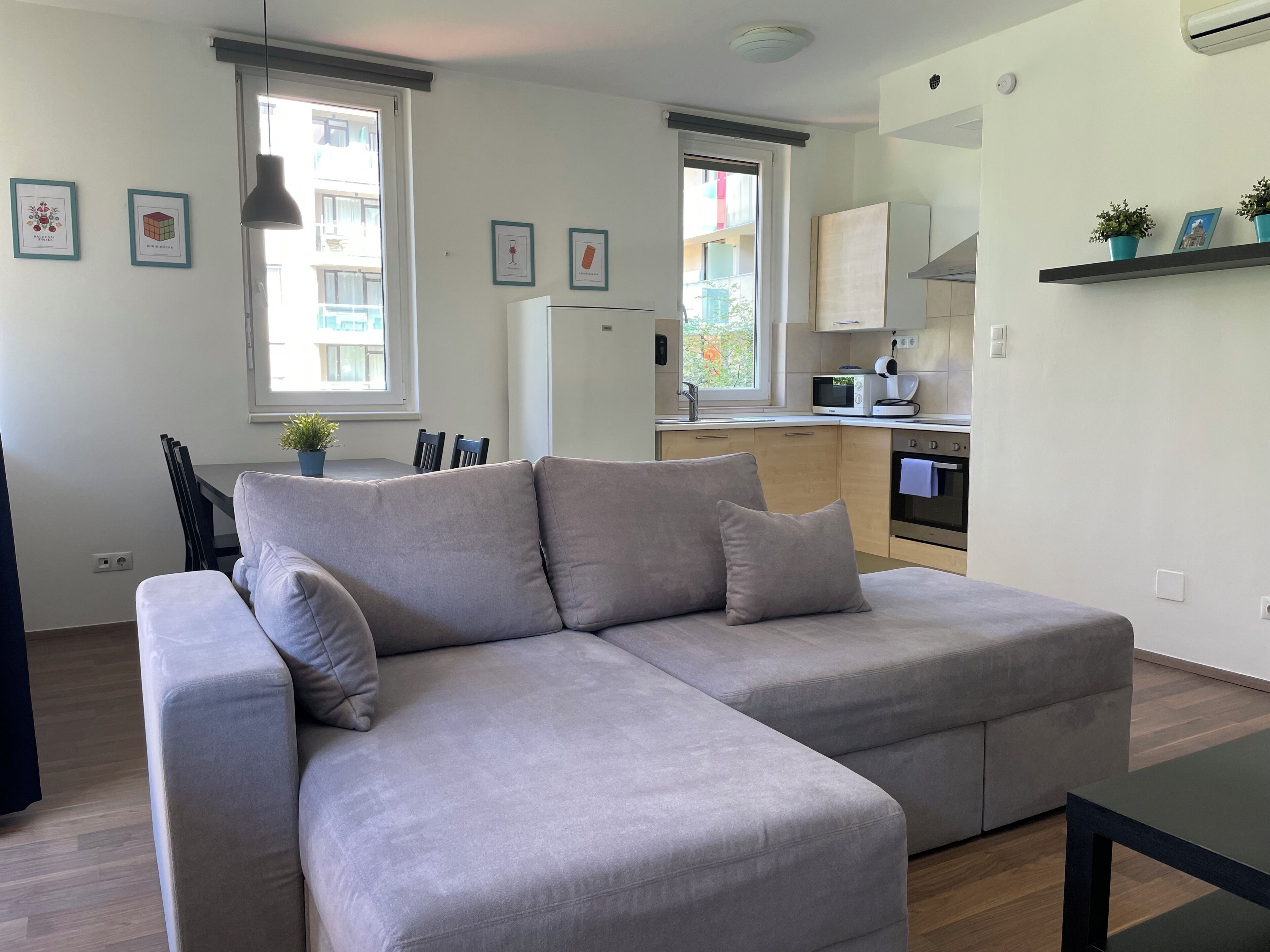 A modern living room with a grey sectional sofa, a dining table with chairs, a kitchen area with appliances, and framed pictures on the walls.