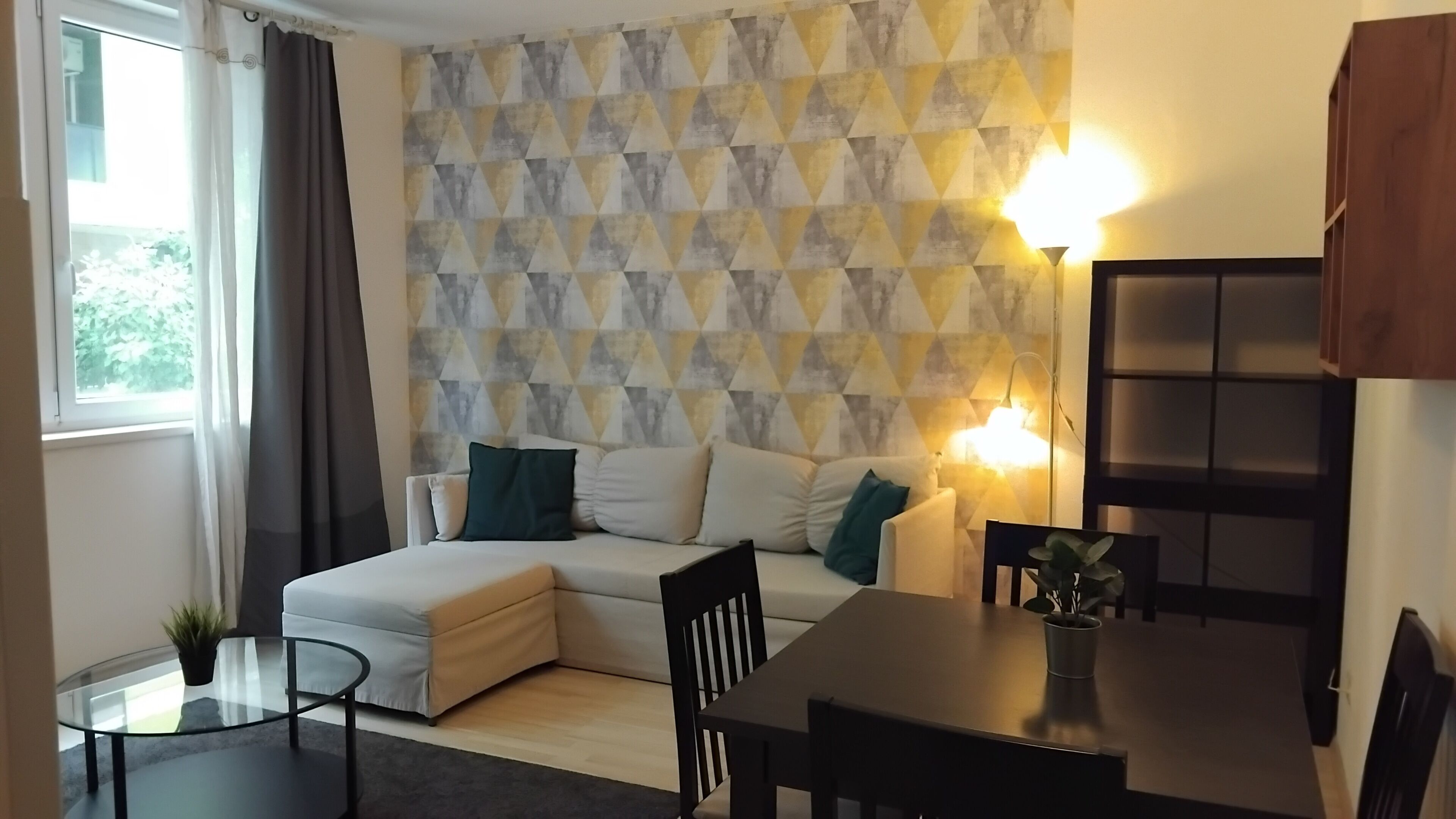 Ein modernes Wohnzimmer mit einem Ecksofa, einem Glastisch, einem Esstisch und einer Wand mit geometrischem Muster.