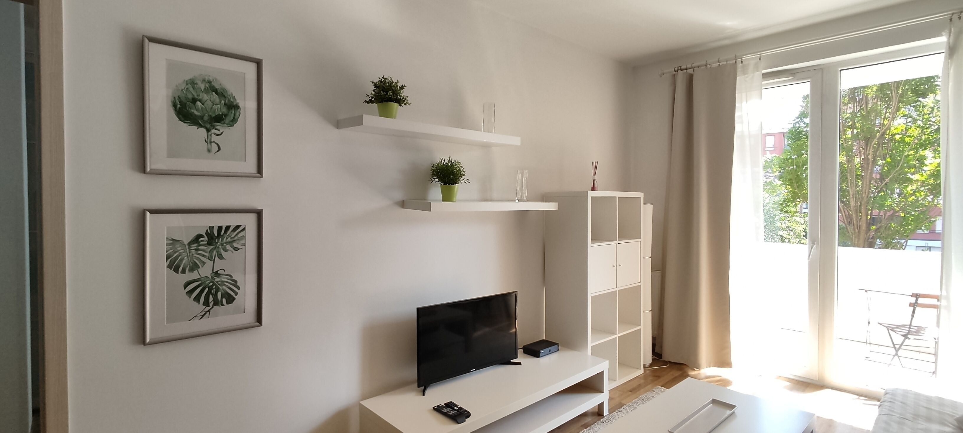Una sala de estar moderna con un televisor de pantalla plana, estanterías blancas con plantas y una puerta corrediza de vidrio al exterior.