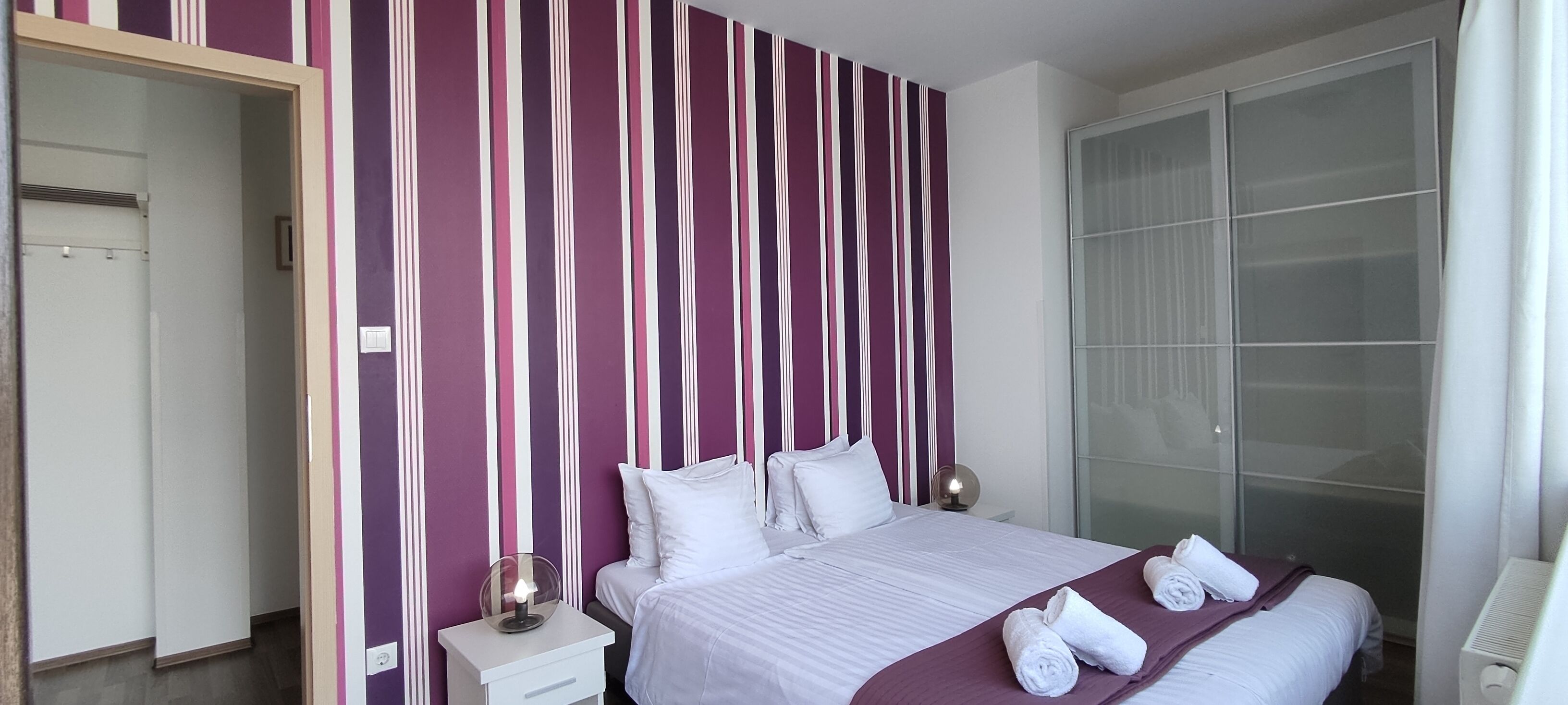 Una camera d'albergo con un letto, biancheria da letto bianca, un comodino con una lampada, un armadio e un'ampia finestra.