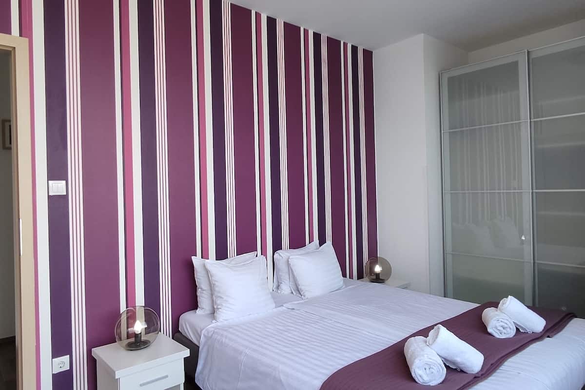 A hotel room with a bed, white bedding, a nightstand with a lamp, a wardrobe, and a large window.