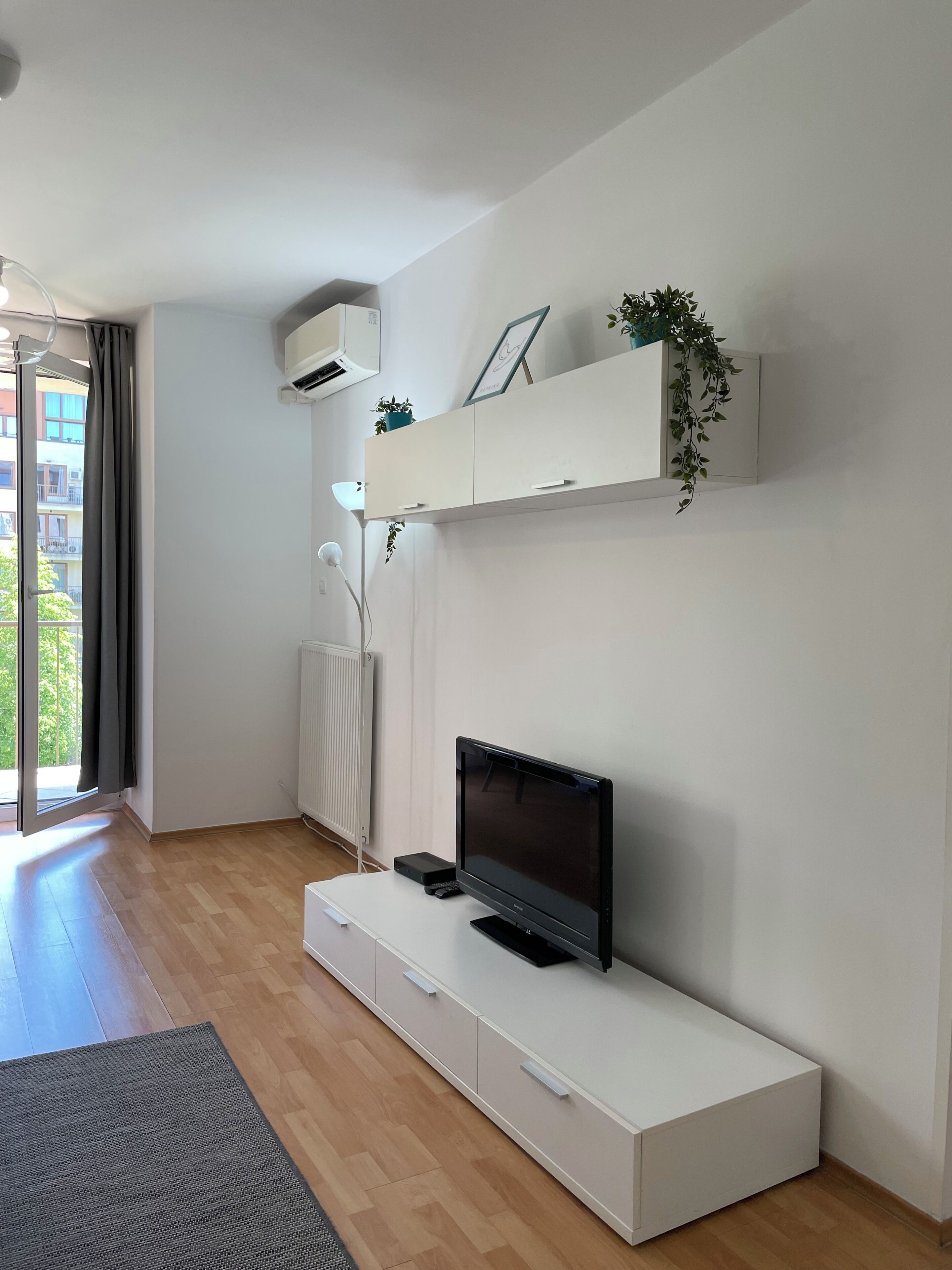 Una sala de estar moderna con un televisor de pantalla plana sobre una consola blanca, un radiador y una planta en maceta en un estante.