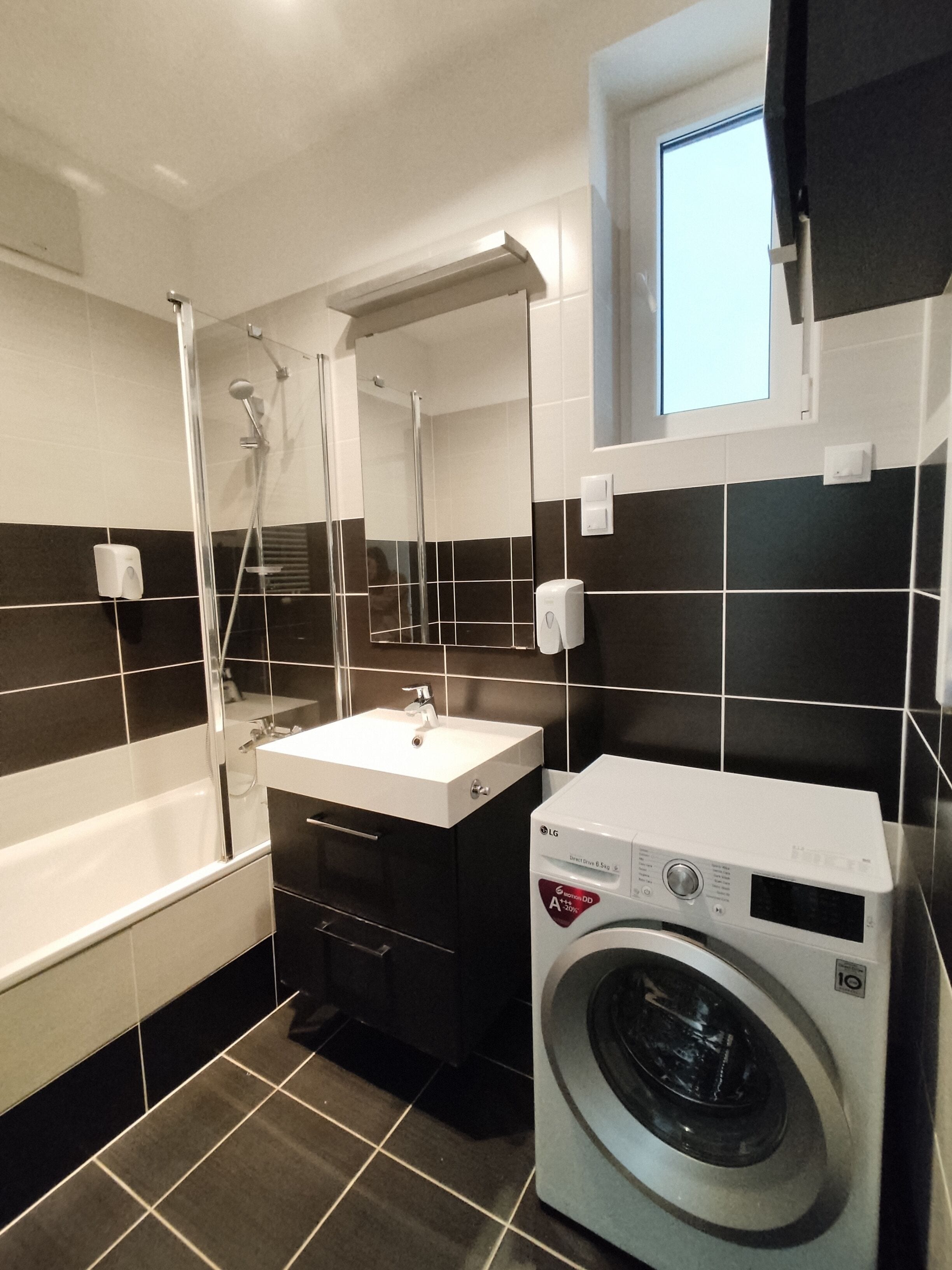A modern bathroom with a white sink, black and white tiles, a glass-enclosed shower, and a front-loading washing machine.