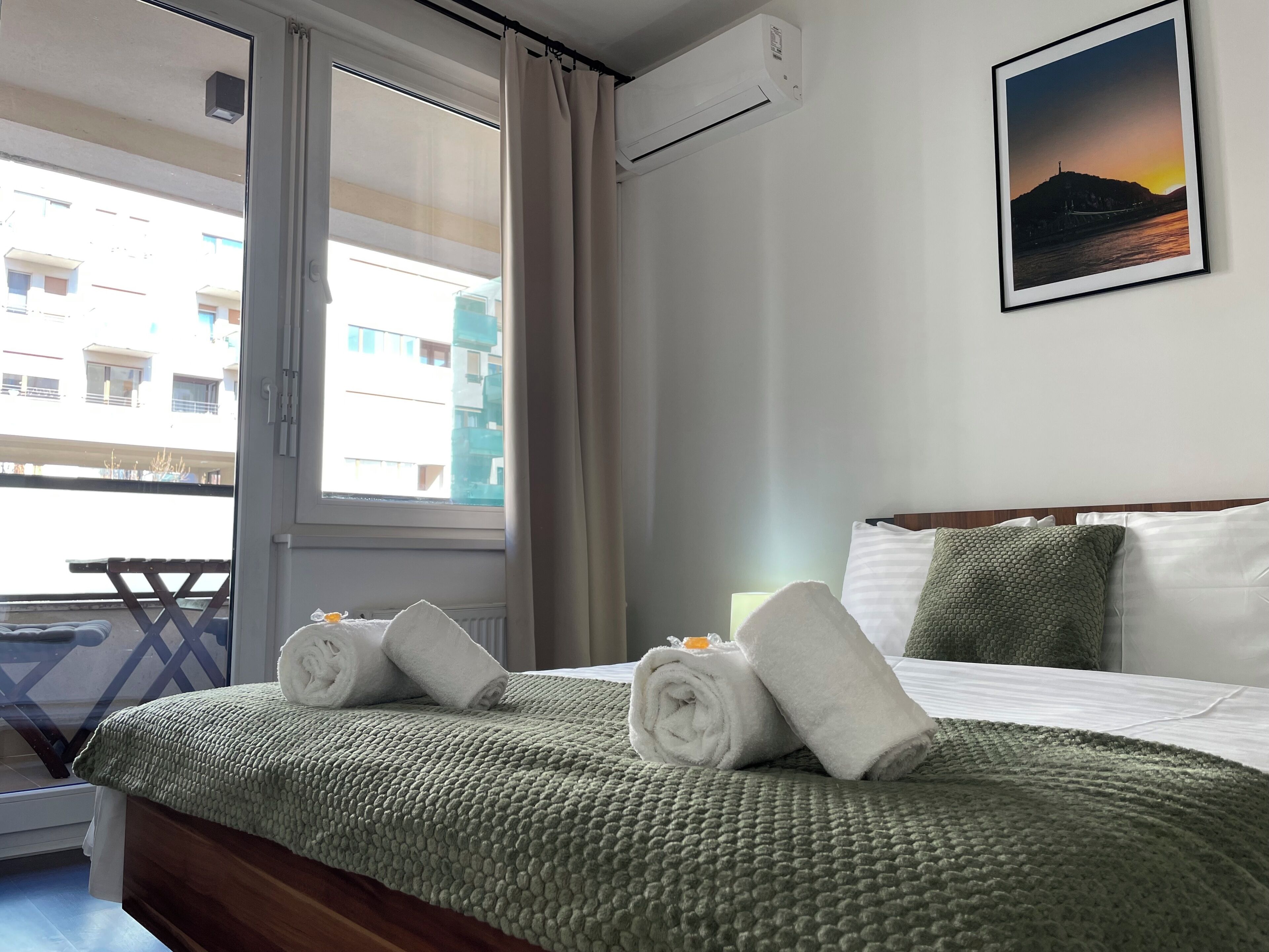 Habitación de hotel con cama, ventana, vista a edificios y un cuadro en la pared.