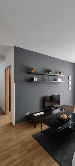 A modern living room with a dark grey wall, wooden flooring, a black coffee table, a flat-screen TV, and two shelves with small plants.