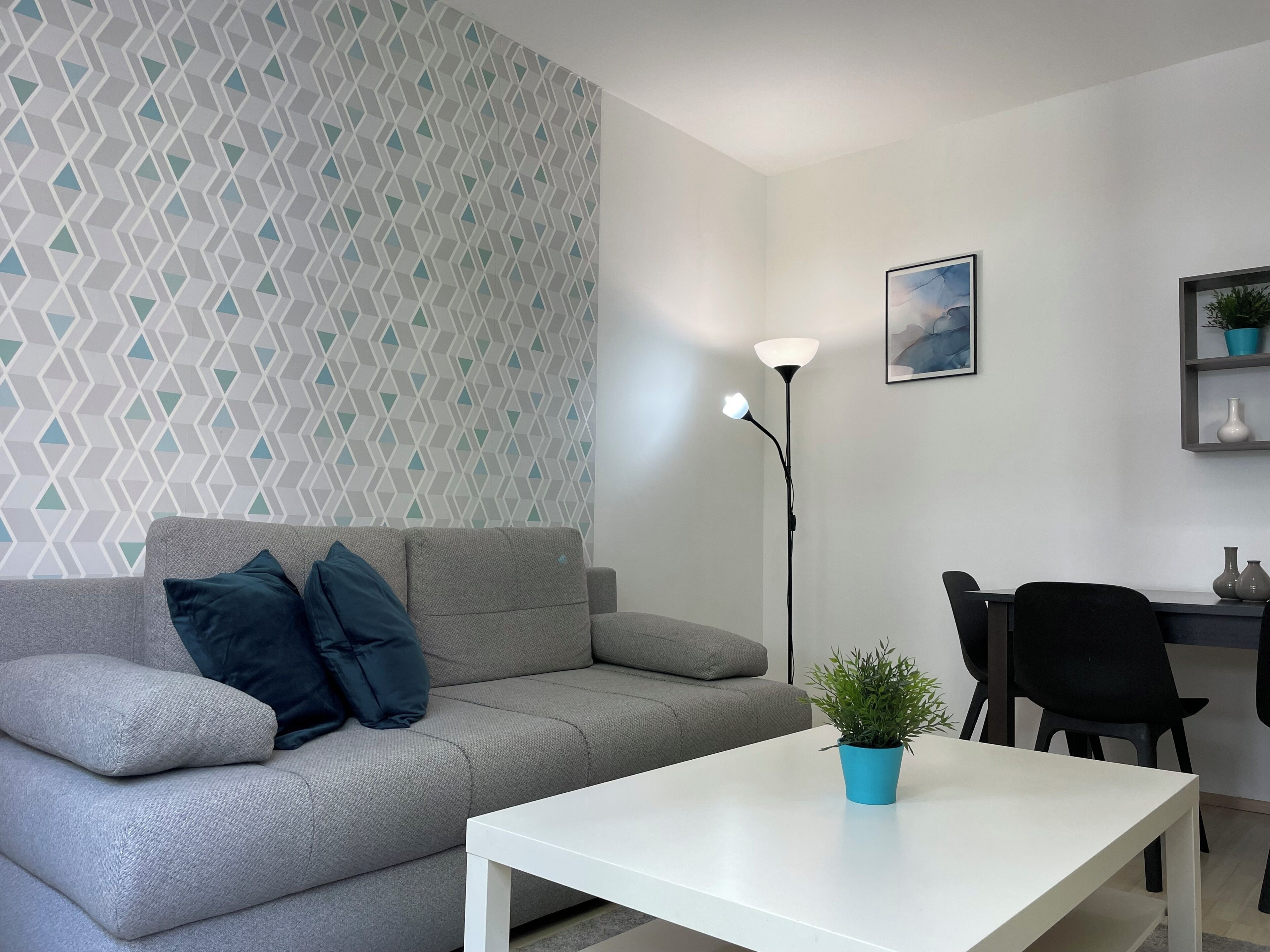 Un salon moderne avec un canapé gris, une table basse blanche, un lampadaire, une plante bleue en pot et un coin repas avec des chaises et une table.