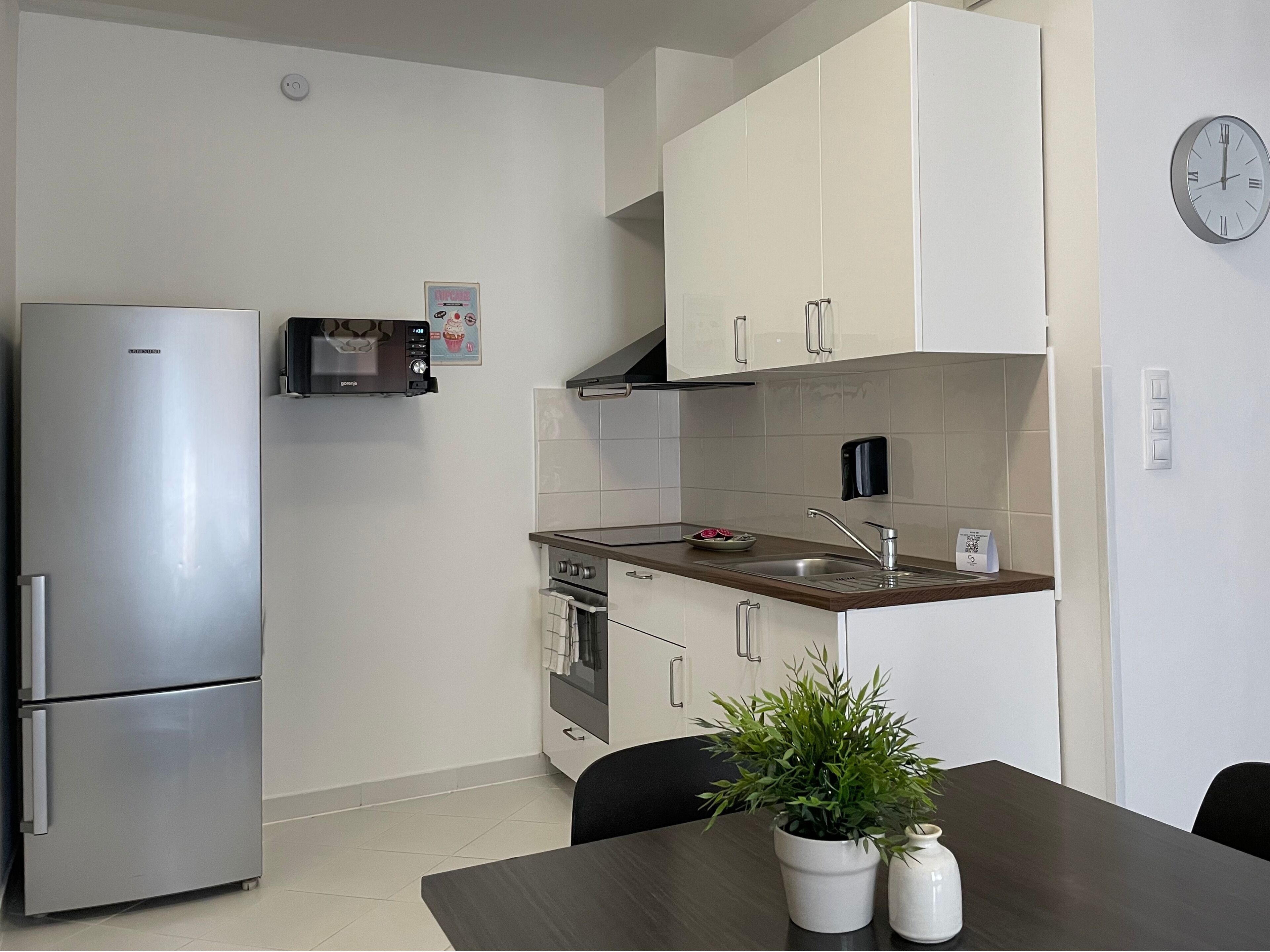 Cocina compacta equipada con refrigerador, microondas, horno y pileta con grifo. Hay un reloj en la pared y una pequeña planta en la mesa.