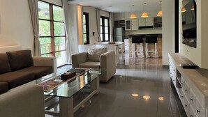 A modern living room with a glass coffee table, sofas, and a kitchen area with pendant lights. - Labak River Hotel by EPS (Ubud)