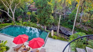 A pool area with lounge chairs and red umbrellas, surrounded by lush greenery and a view of a traditional house. - Labak River Hotel by EPS (Ubud)
