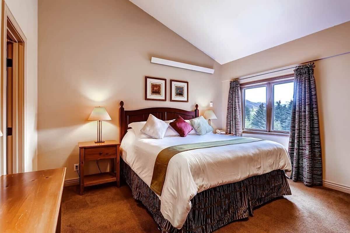 A bedroom with a large bed, two bedside tables, a window with curtains, and two framed pictures on the wall.