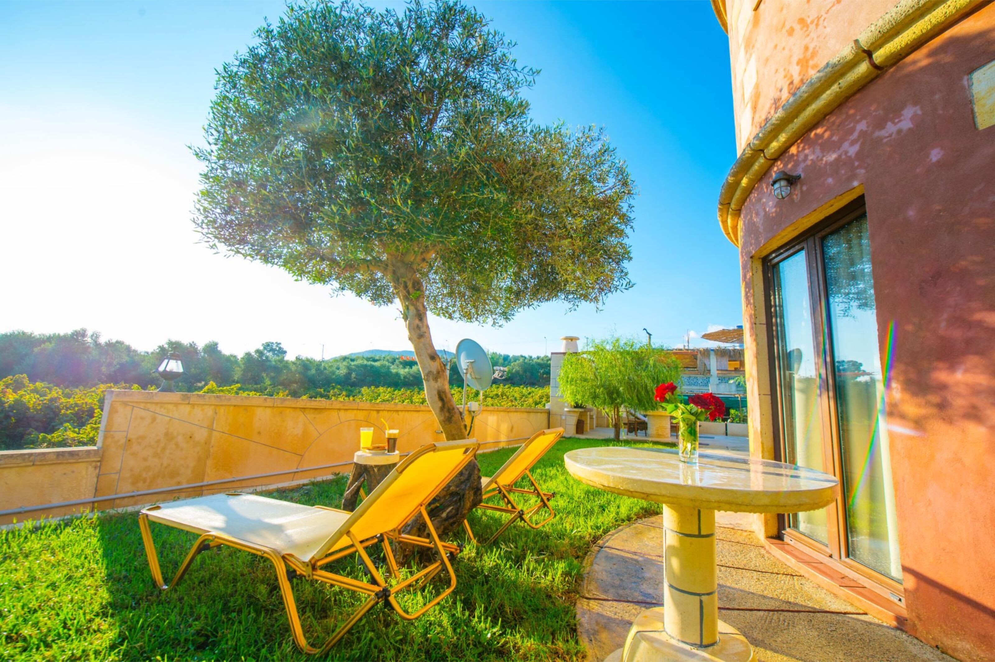 A terrace with two yellow lounge chairs and a table, overlooking a lush landscape and a building with large windows.