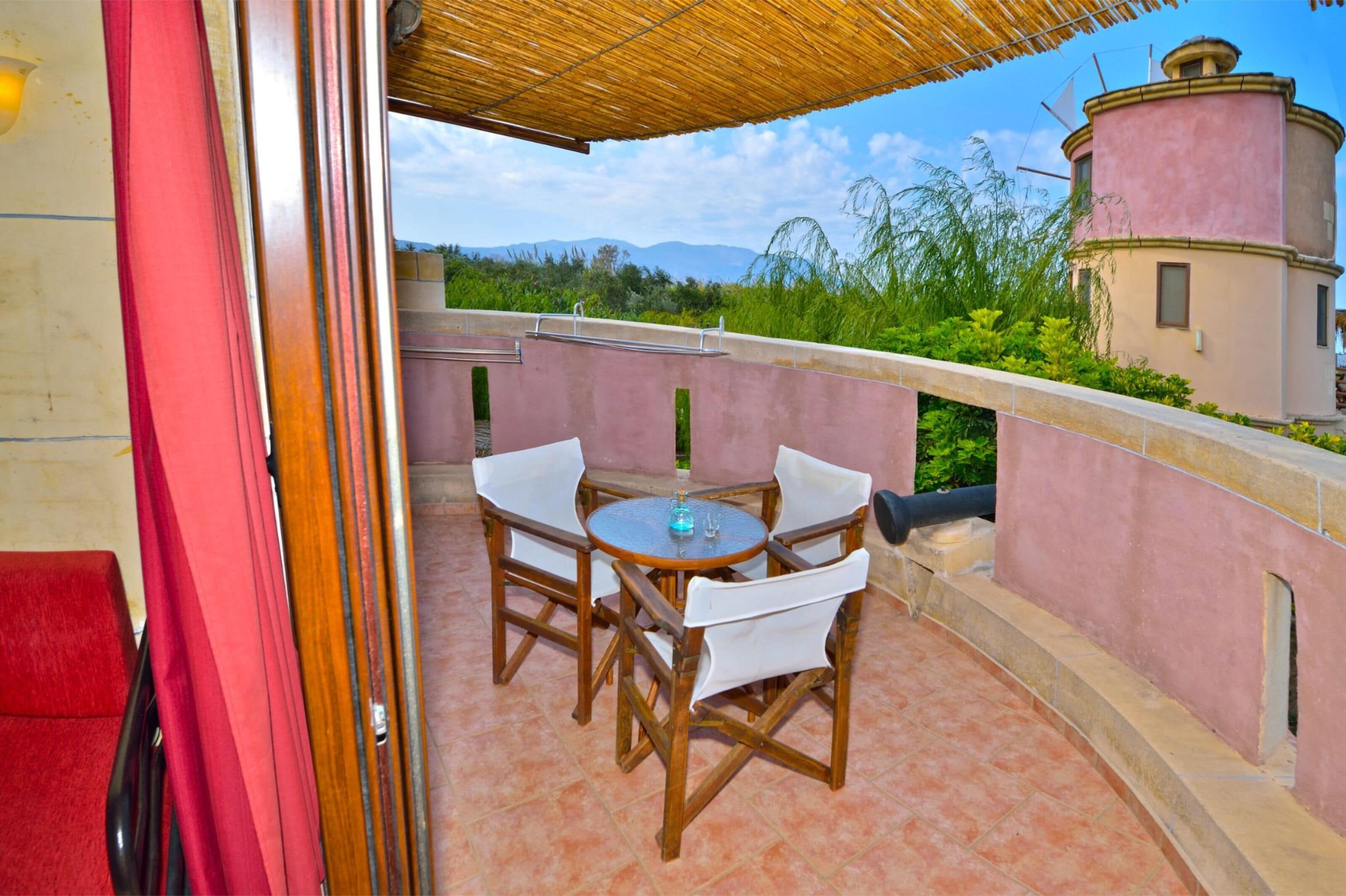 Uma varanda com mesa e cadeiras, vista para área verde e montanhas, e um edifício rosa ao fundo.