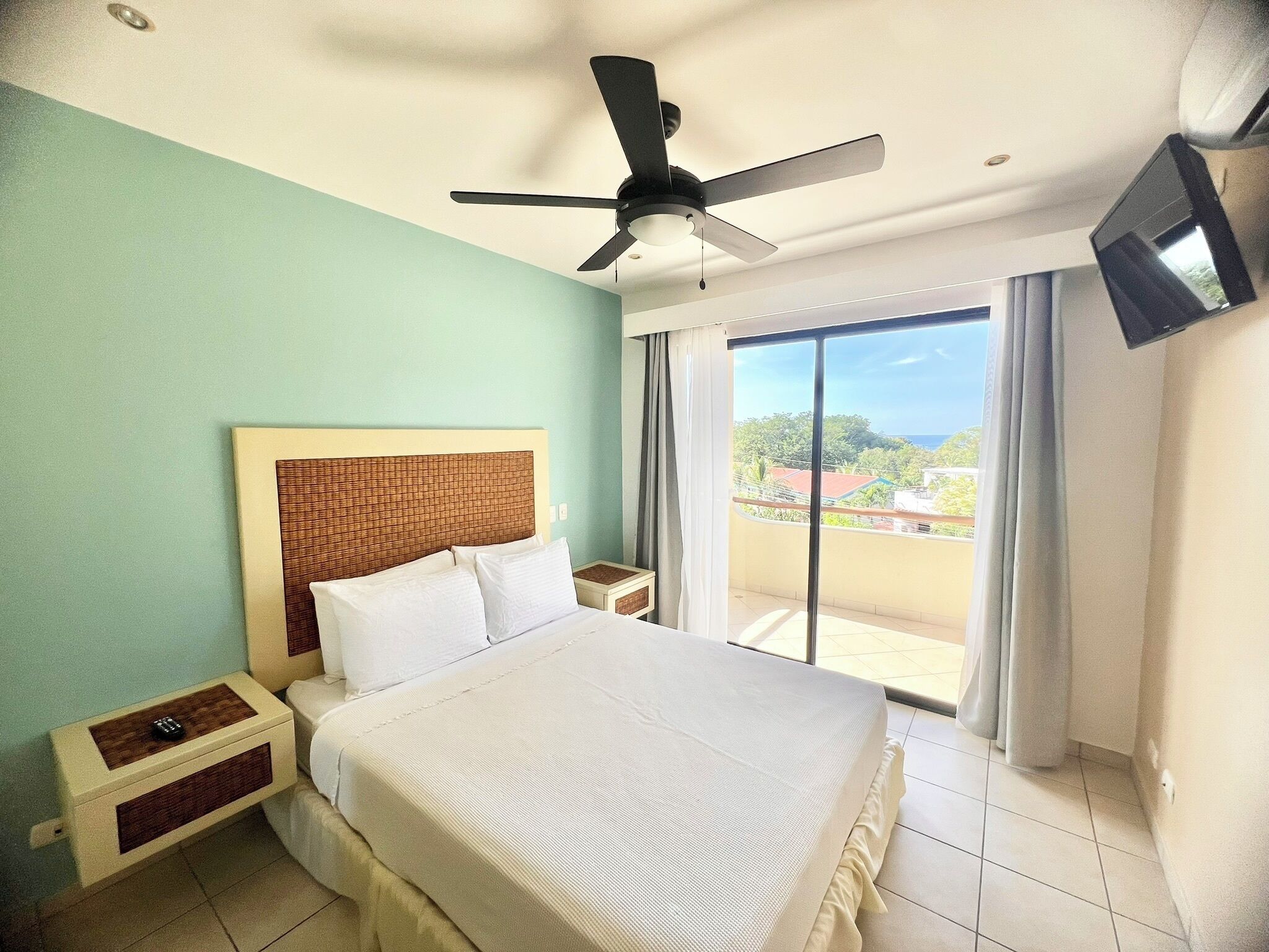 A hotel room with a bed, a ceiling fan, a TV mounted on the wall, and a view of the outside through a large window.