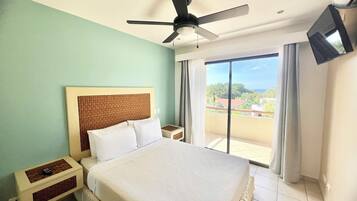 A hotel room with a bed, a ceiling fan, a TV mounted on the wall, and a view of the outside through a large window.