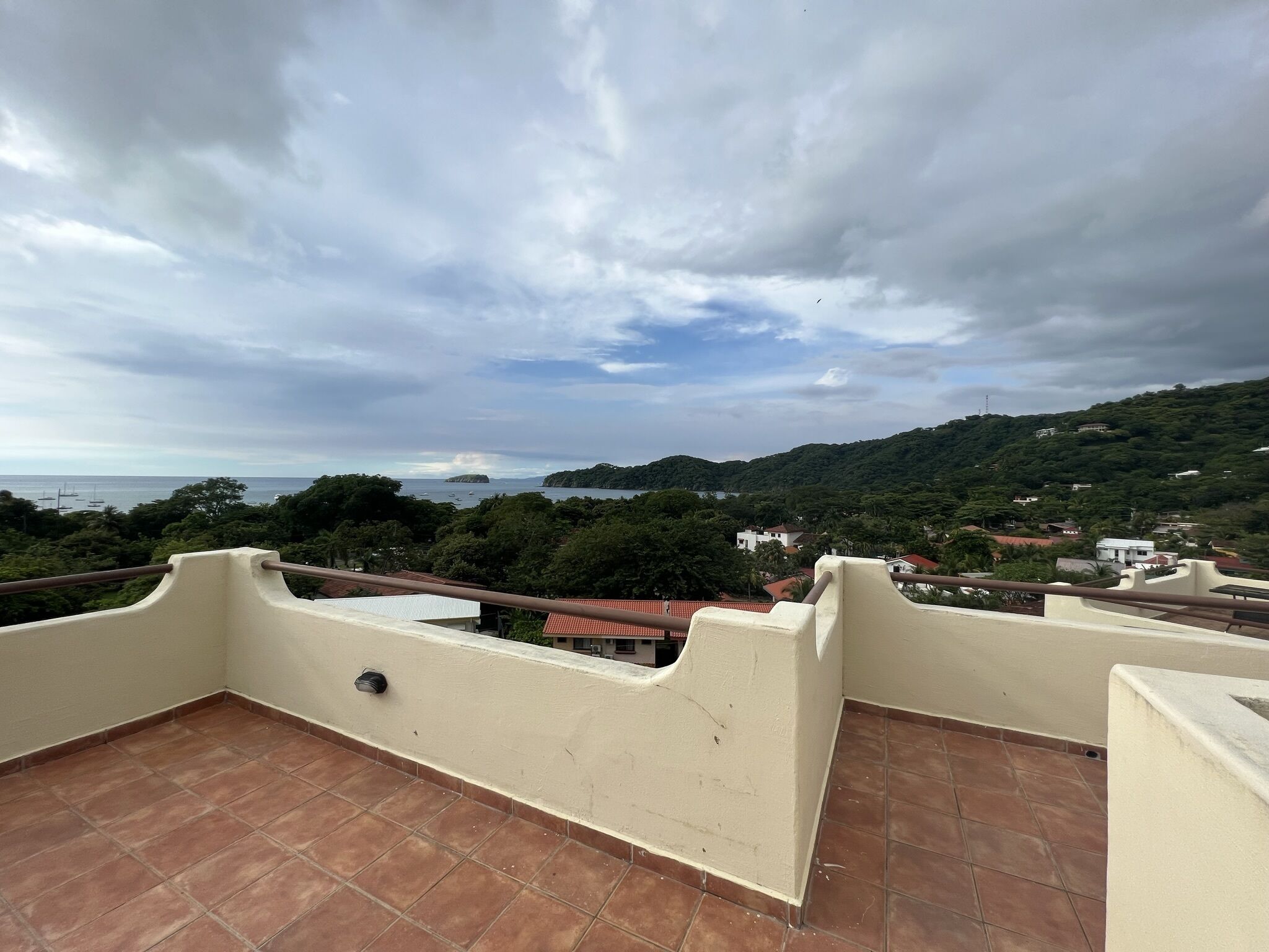 A rooftop terrace with a view of a coastal landscape, including a hill with vegetation and buildings.