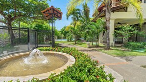 A landscaped garden with a central water feature, surrounded by lush greenery and a pathway leading to a building with a covered entrance. - Tropical Gardens (Sardinal)