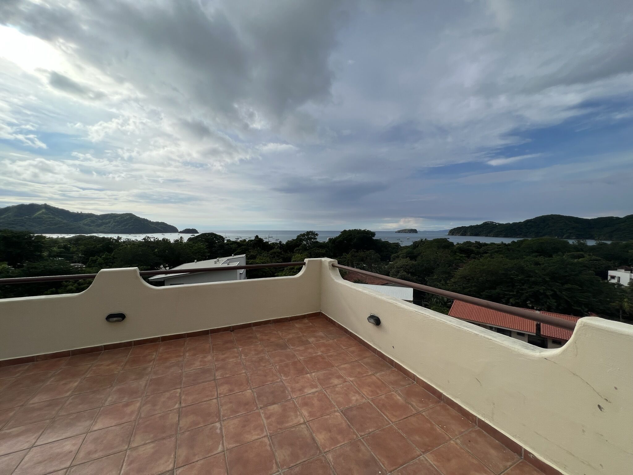 A terrace with a tiled floor and a railing, overlooking a coastal landscape with hills and a body of water.