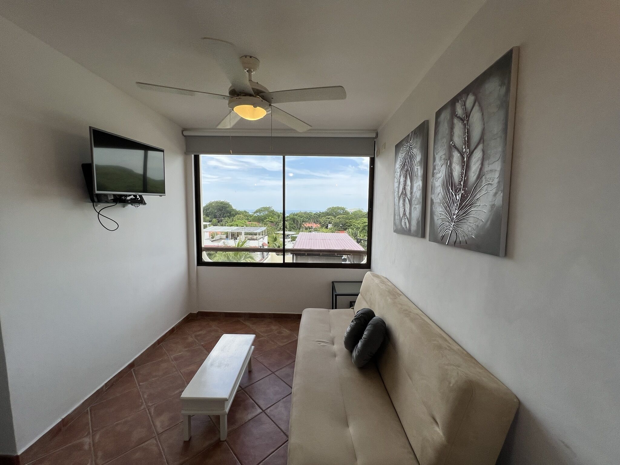 A room with a ceiling fan, a large window with a view of buildings and trees, a flat-screen TV mounted on the wall, and a beige sofa with a pillow.