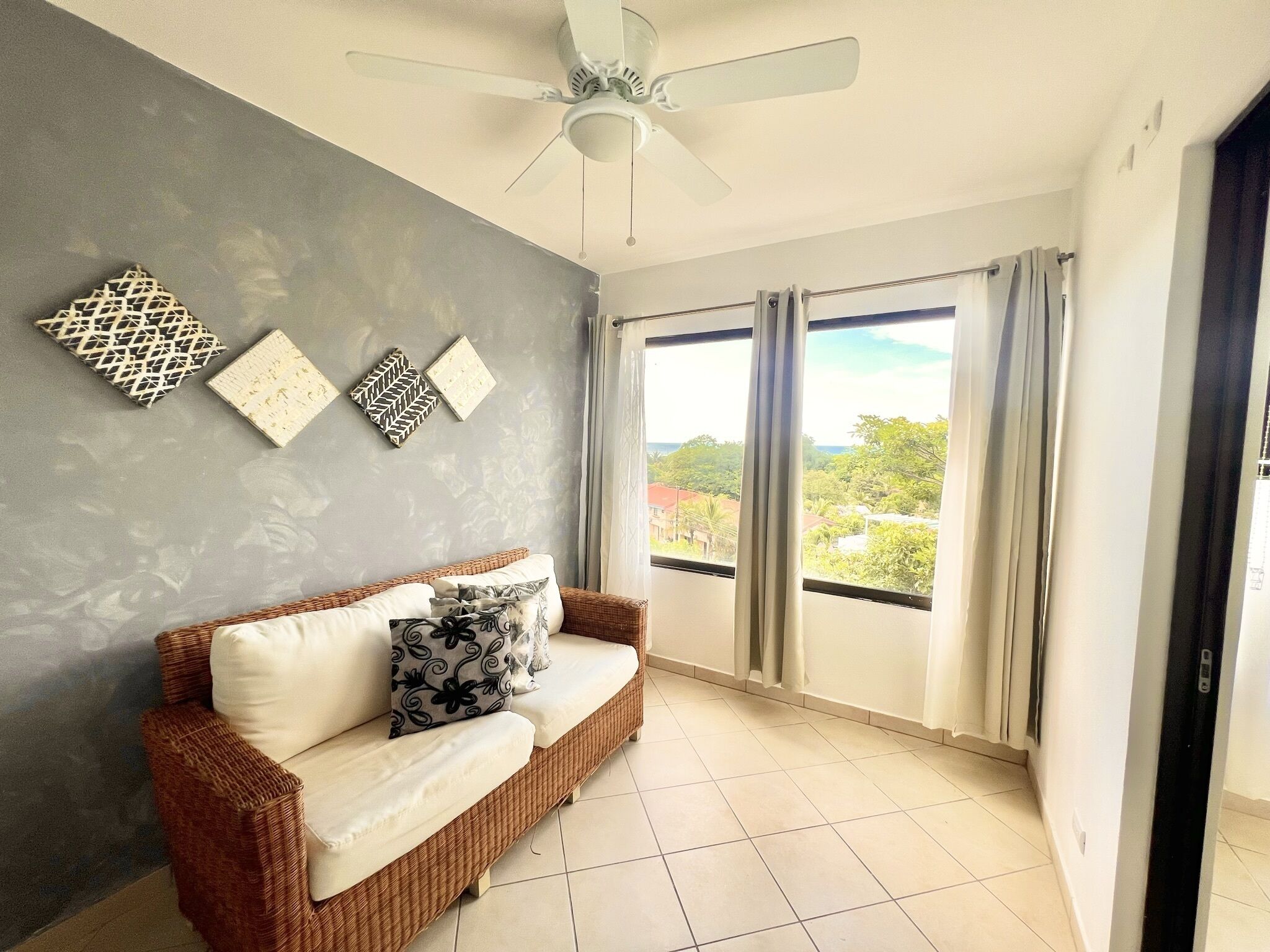 A room with a wicker sofa, a ceiling fan, and a large window with curtains.