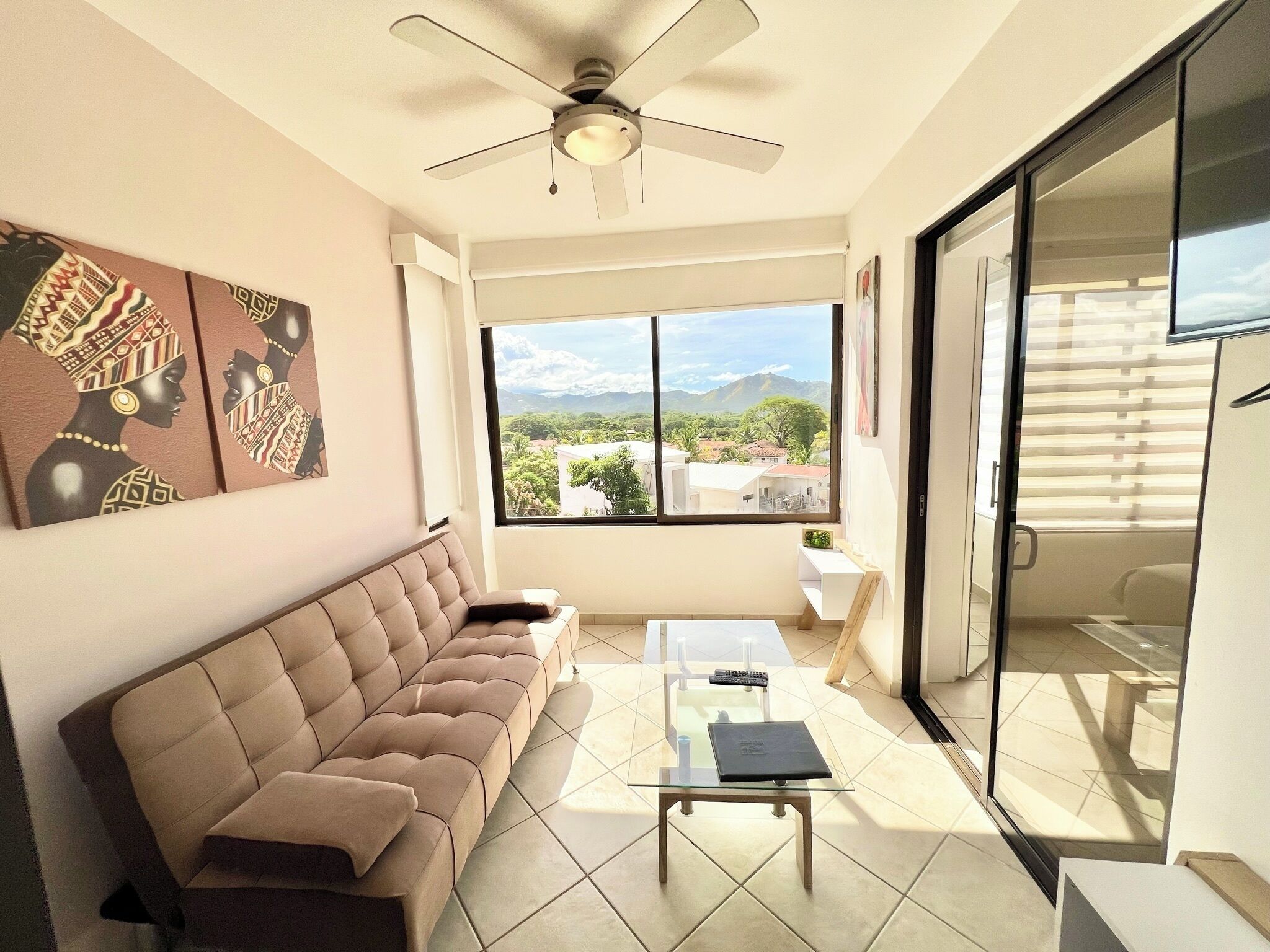 A modern living room with a sofa, a glass coffee table, a ceiling fan, and a large window offering a view of a cityscape.