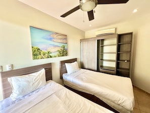 A hotel room with two beds, a ceiling fan, a painting of a beach, and a wardrobe with shelves. - Tropical Gardens (Sardinal)