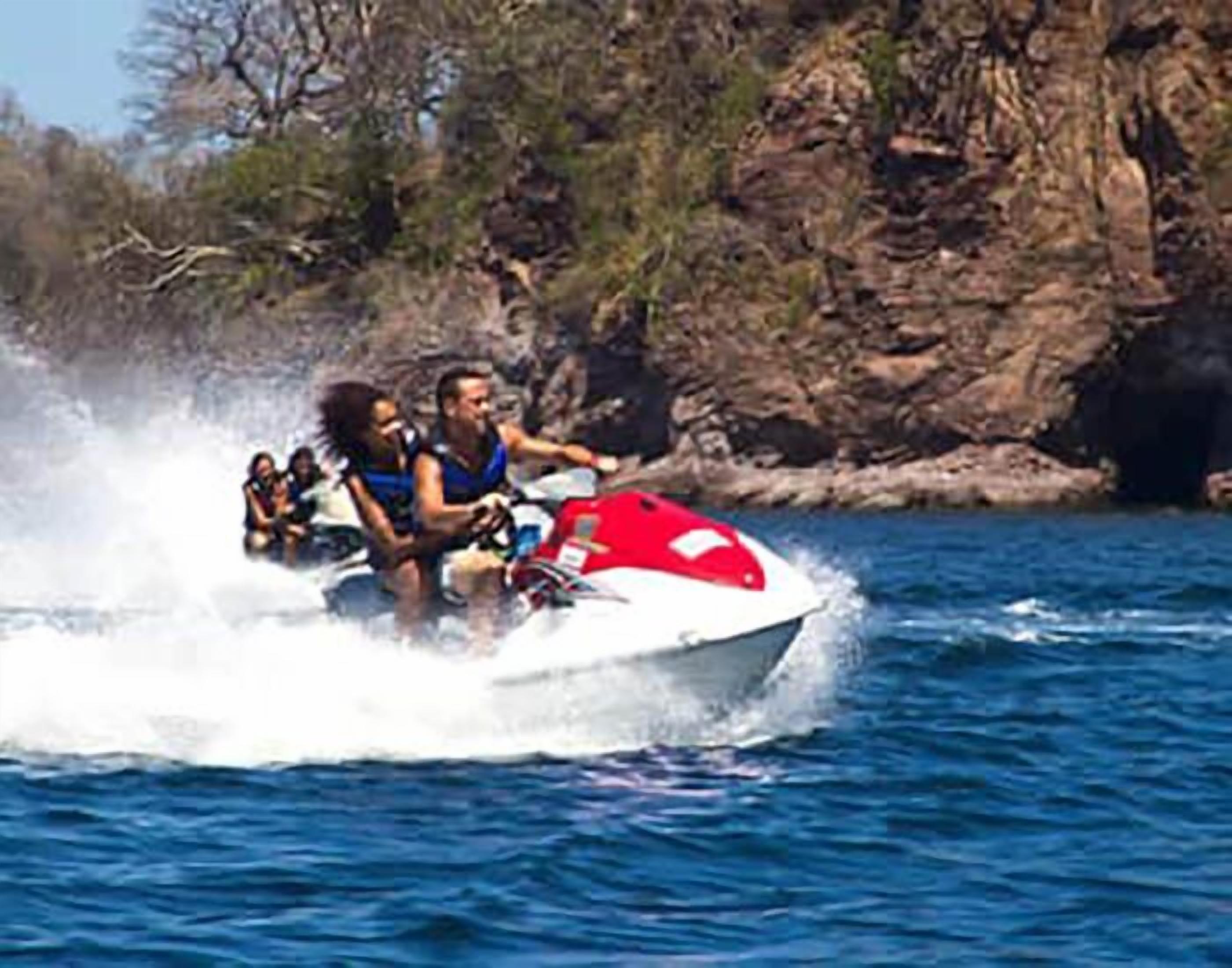 A jet ski with passengers on a body of water, surrounded by rocky terrain and sparse vegetation.