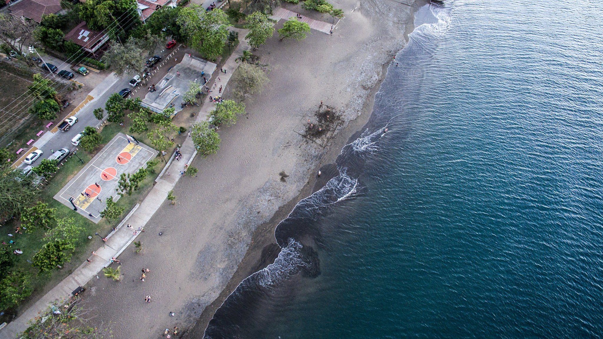 Vista aérea de una playa con canchas de baloncesto, un estacionamiento y una zona residencial junto a la costa.
