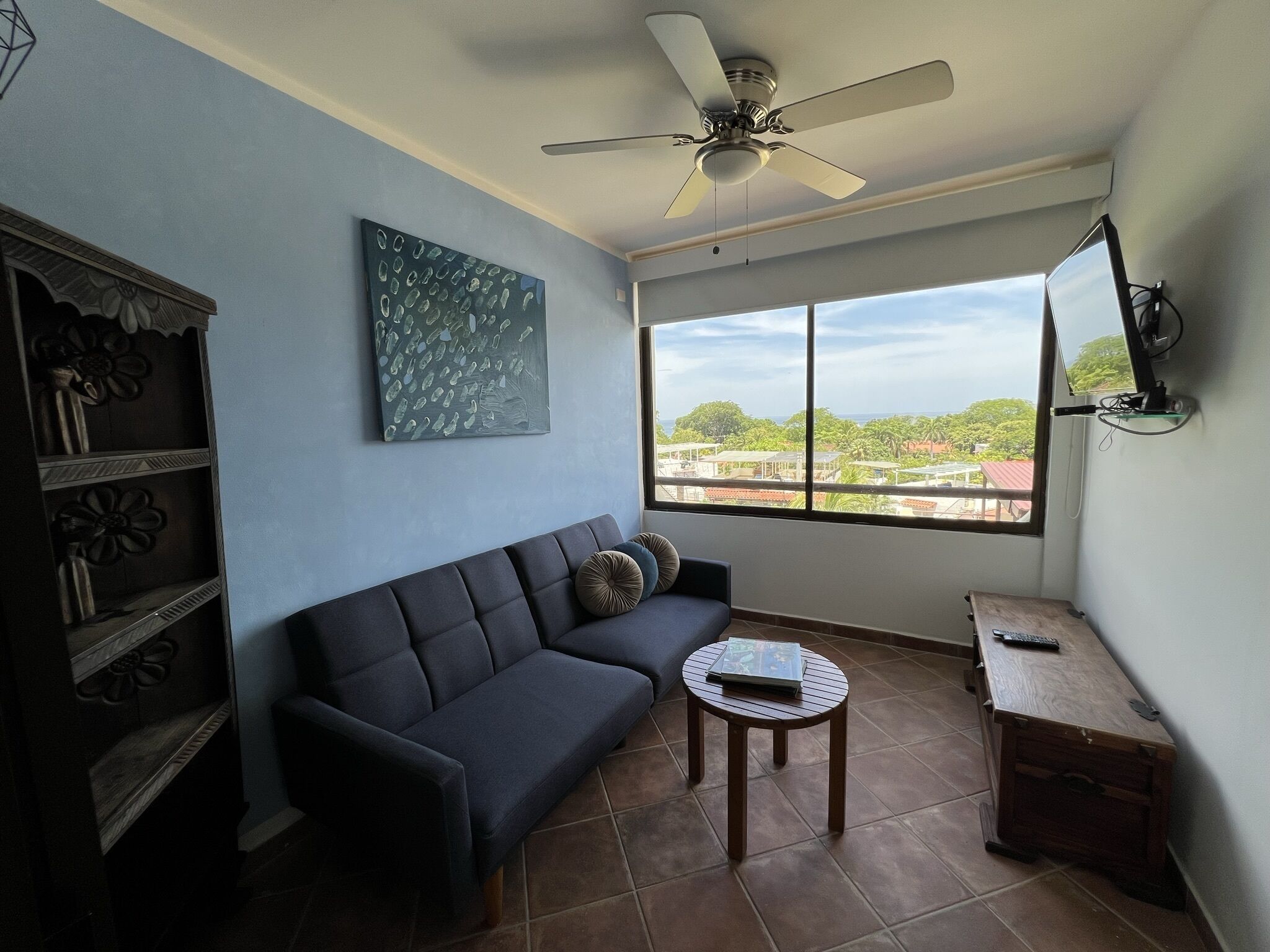 A living room with a dark sofa, a small round table, a ceiling fan, a large window with a view, and a decorative cabinet.