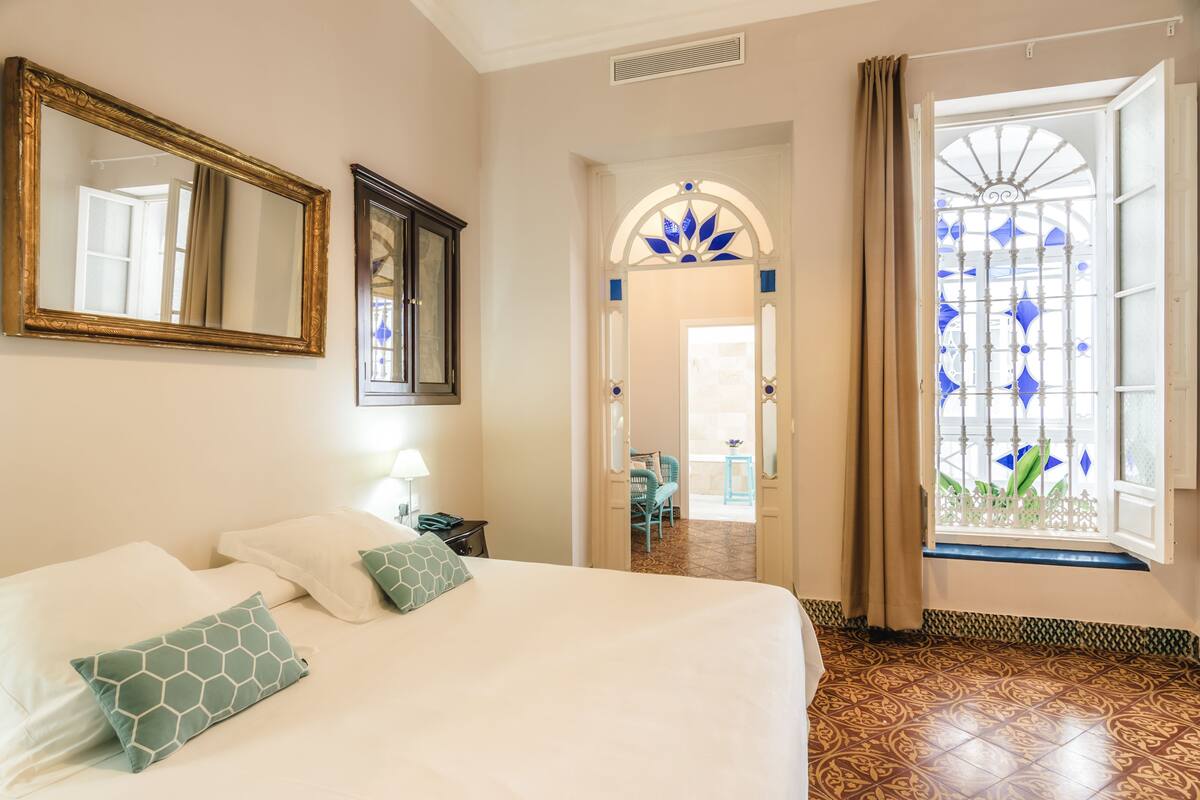 Ein Hotelzimmer mit einem großen Bett, einem Spiegel, einem Schreibtisch mit Lampe und Blick durch ein Fenster mit einer verzierten Glasschiebetür.