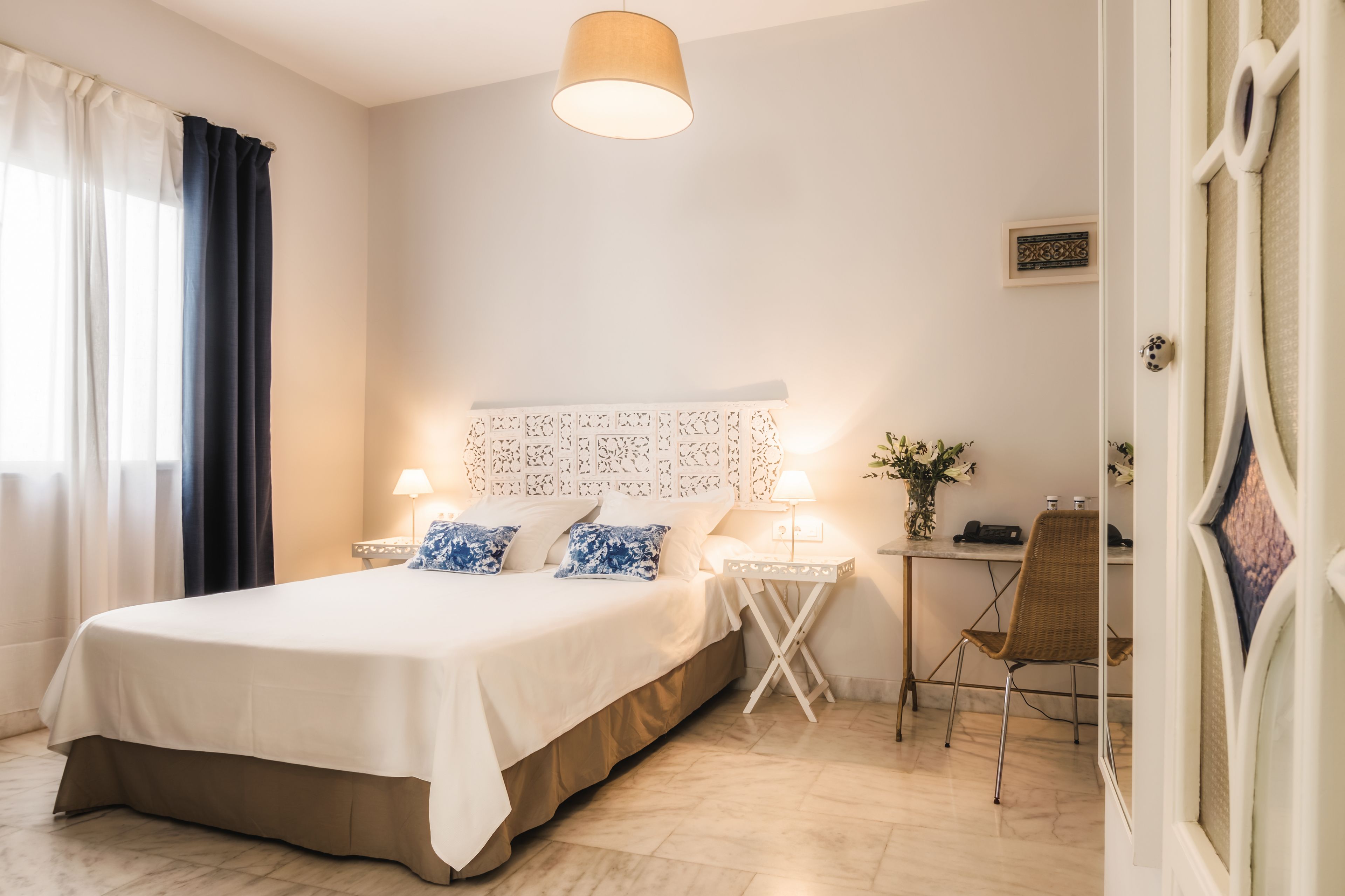 A neatly arranged hotel room with a bed, bedside lamps, a small table with a chair, and a decorative headboard.