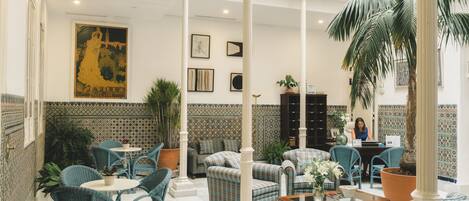 A spacious lobby with high ceilings, a central seating area with chairs and sofas, a large potted plant, and a decorative wall with a poster.