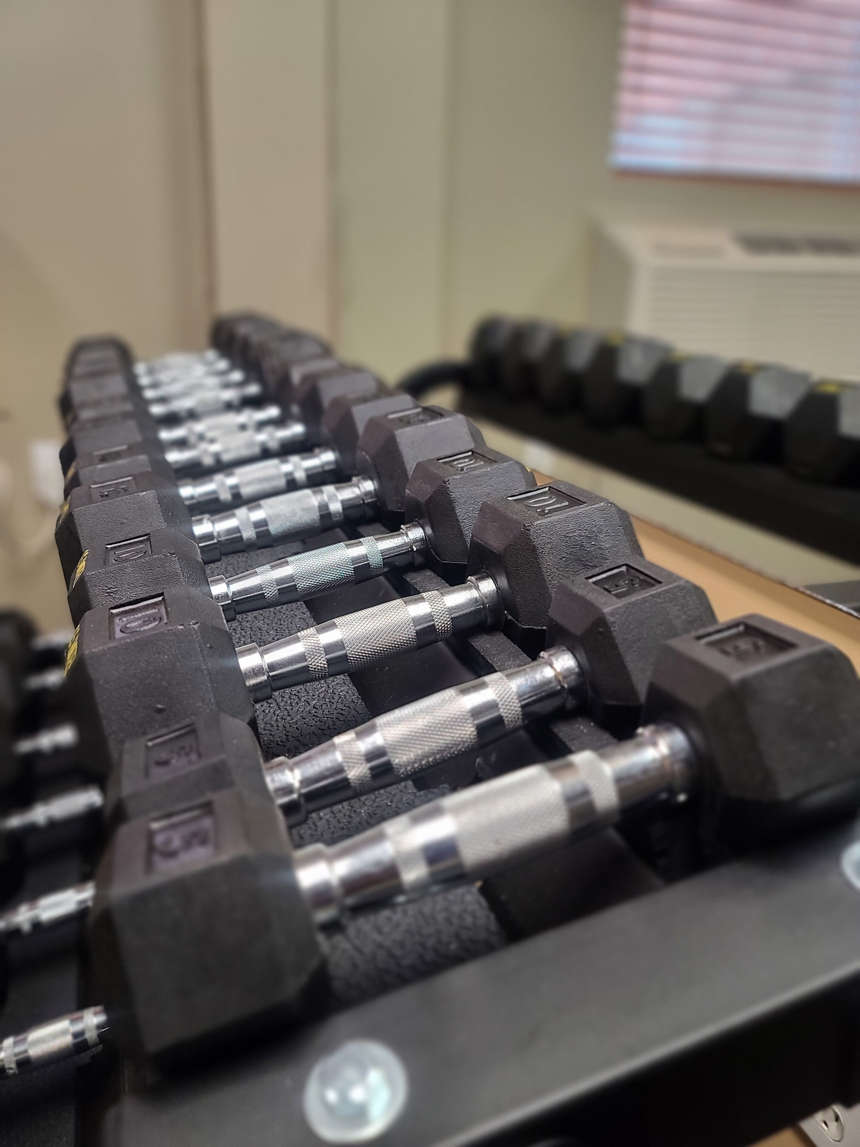 A rack of black dumbbells with adjustable weights, a cable, and a wall-mounted computer monitor in the background.