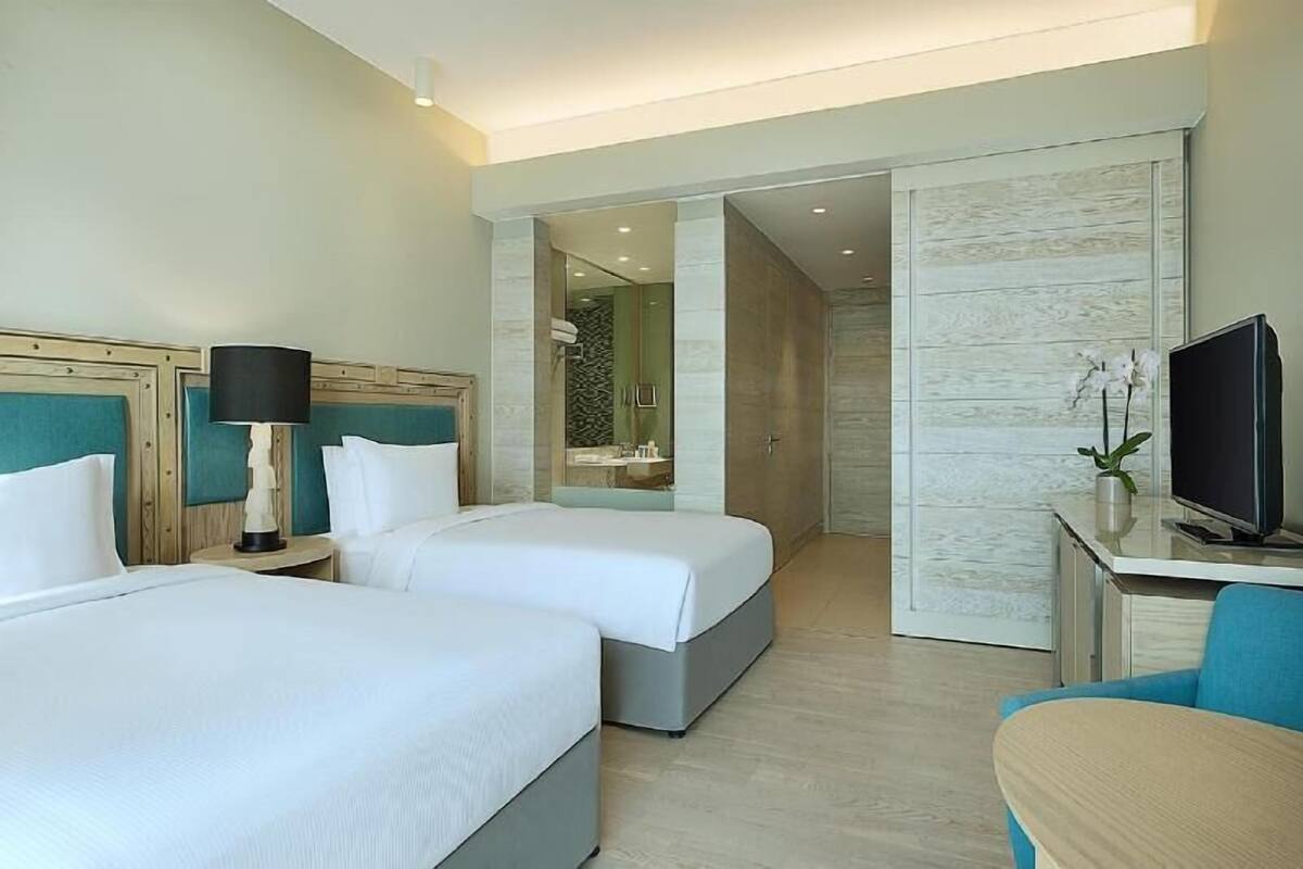 A hotel room with two beds, a desk with a TV, a bathroom with a shower, and a view of the outside.