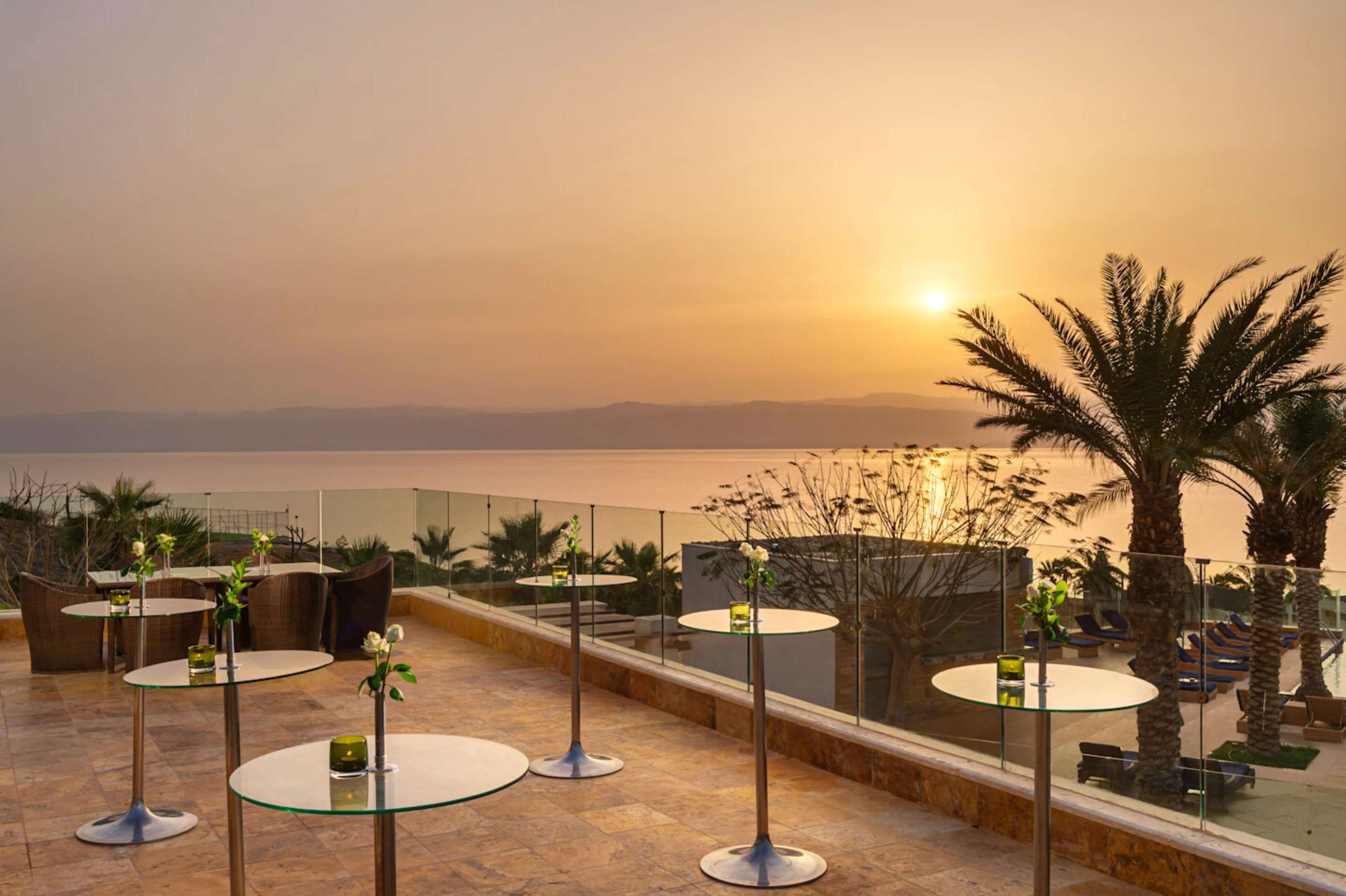 A terrace with round tables and chairs, overlooking a body of water and palm trees.