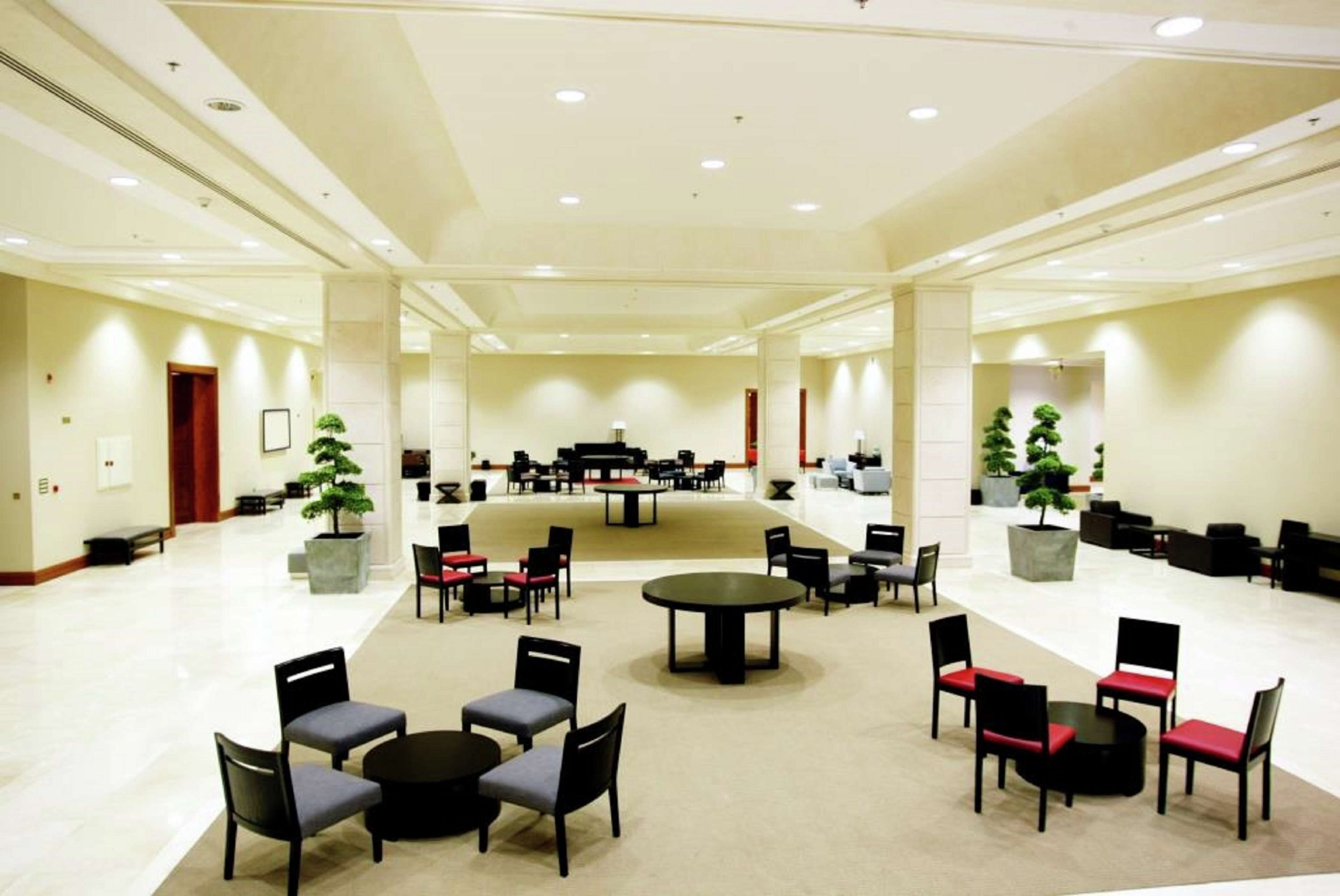 A spacious lobby with multiple seating areas, including round tables and chairs, and several potted plants.