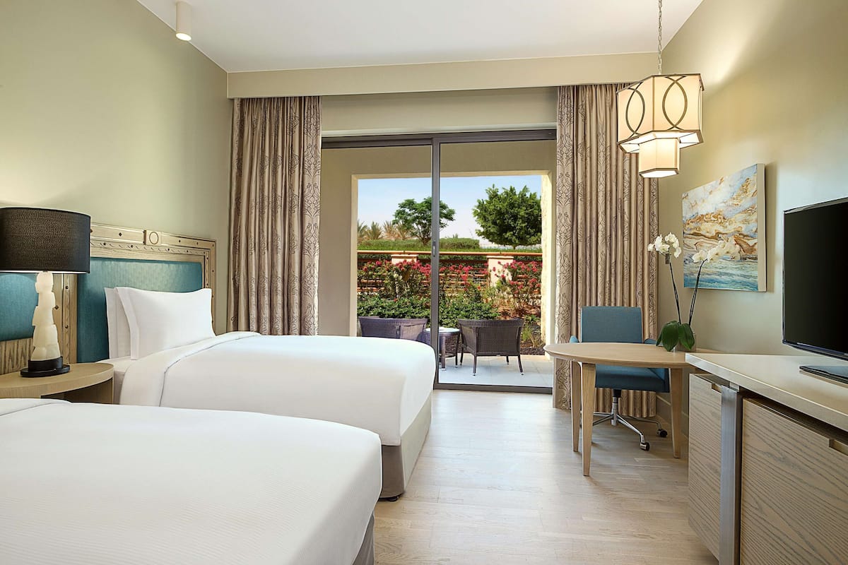 A hotel room with two beds, a desk, a TV, and a view of a garden through a sliding door.