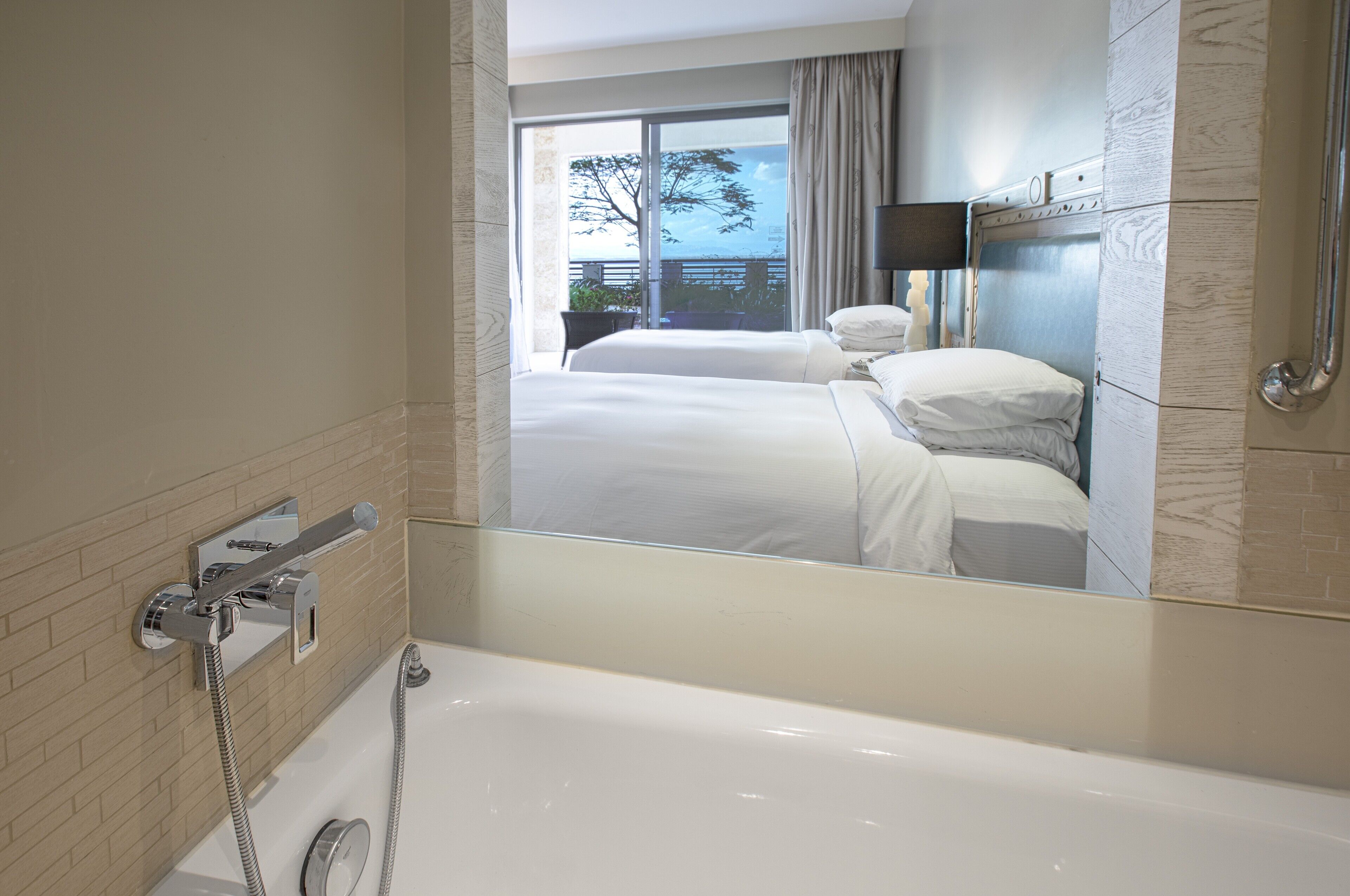 A hotel bathroom with a bathtub, a shower, and a large mirror reflecting a balcony with a view of the sea.