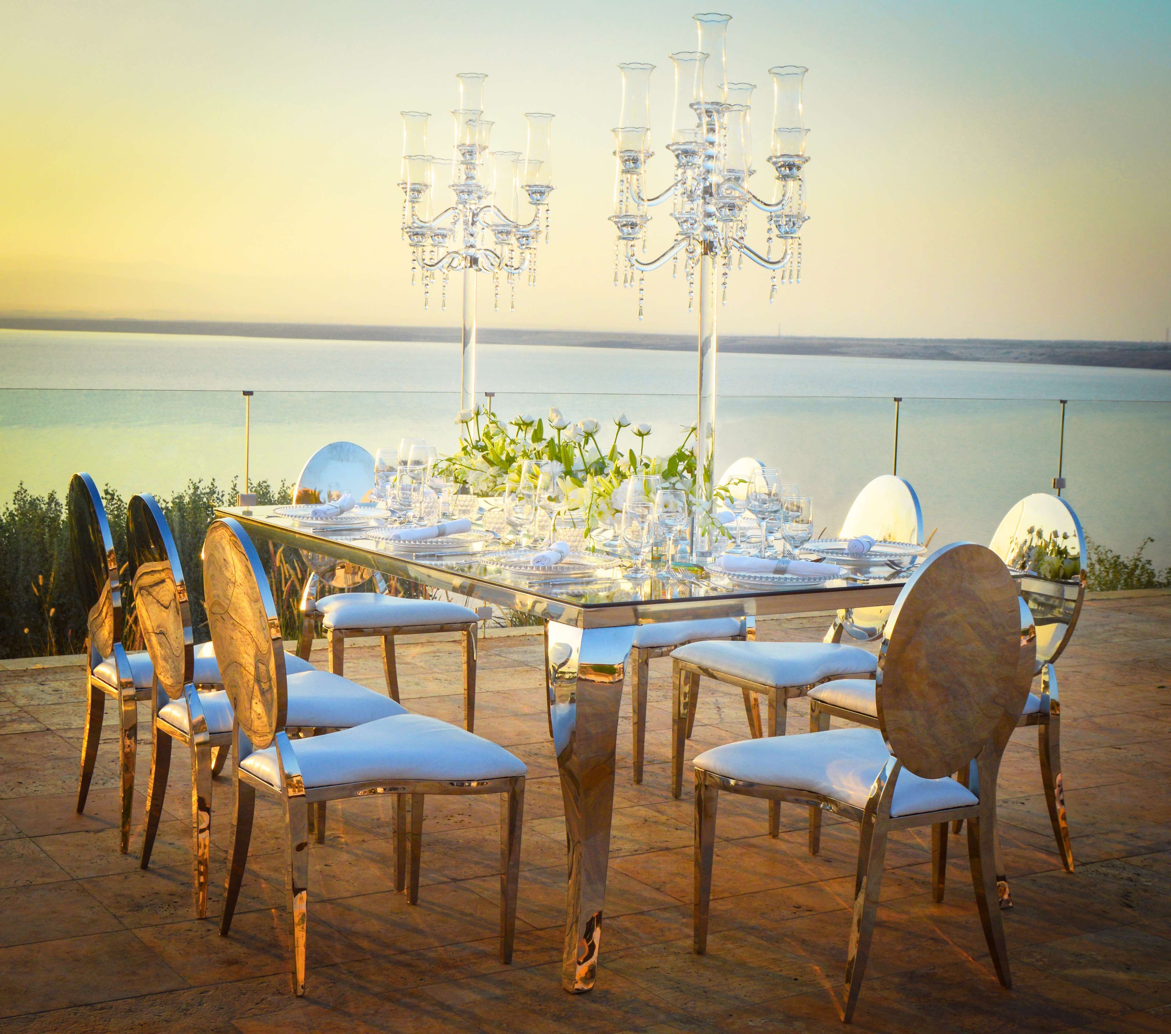An outdoor dining setup with a table set for six, surrounded by chairs, and a chandelier above.
