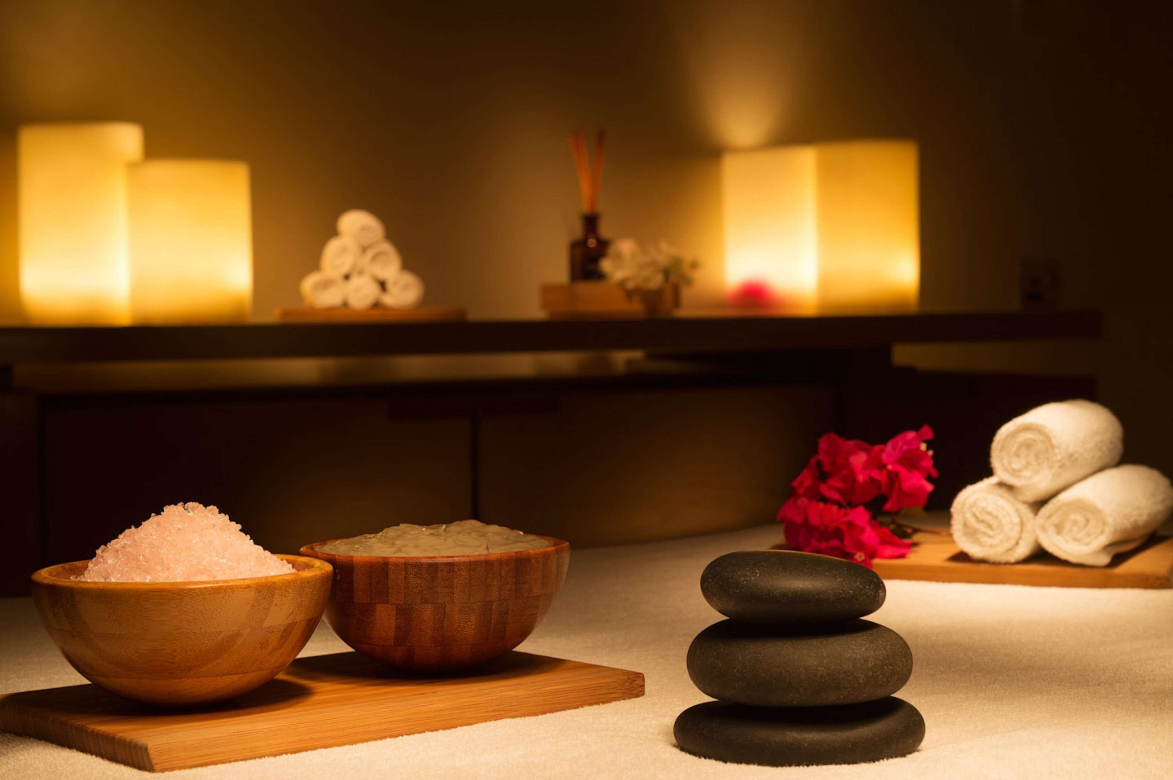 A spa setting with wooden bowls of salts, a stack of stones, and rolled towels on a wooden board.