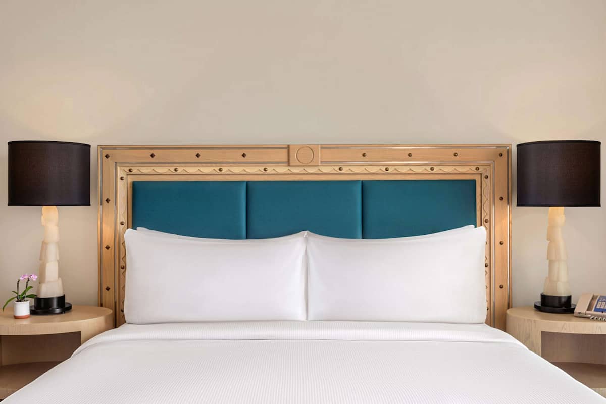 A hotel room with a large bed, two bedside tables, a nightstand, a decorative wall panel, and a potted plant.