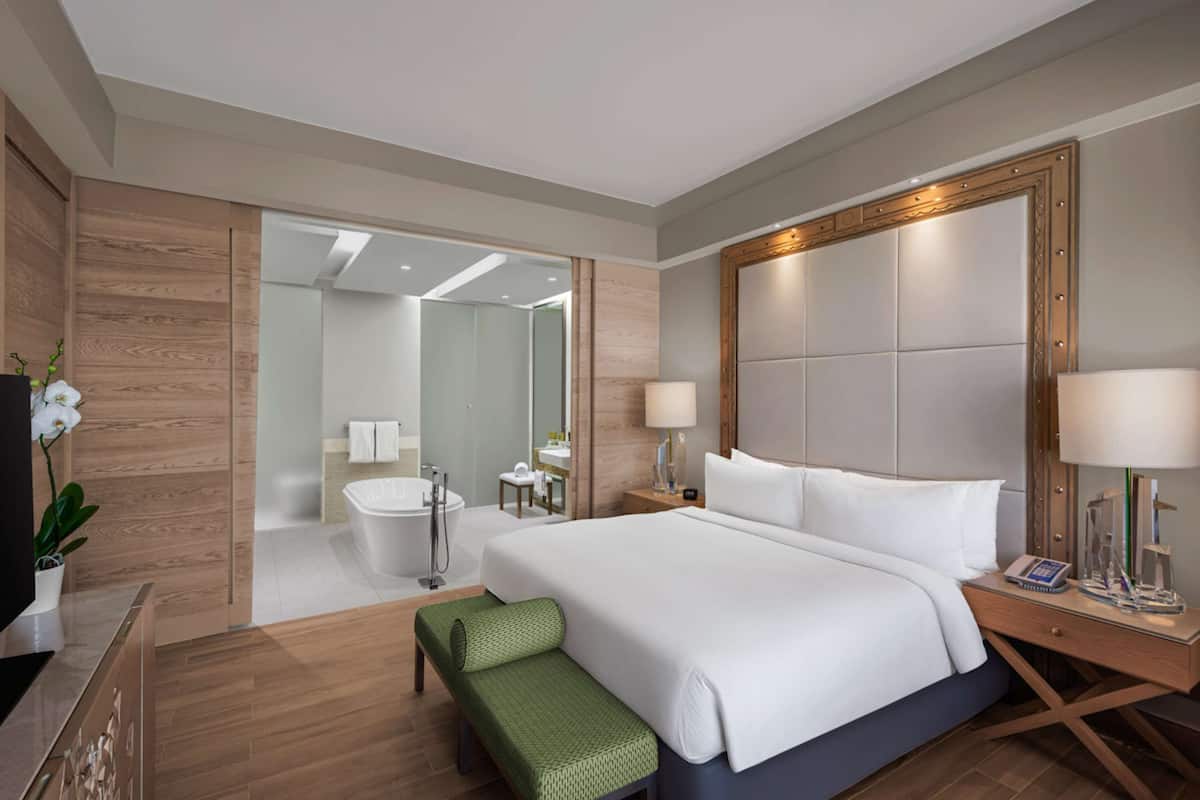 A modern hotel room with a large bed, a green bench, a bedside table with a lamp, and a bathroom visible through an open door.